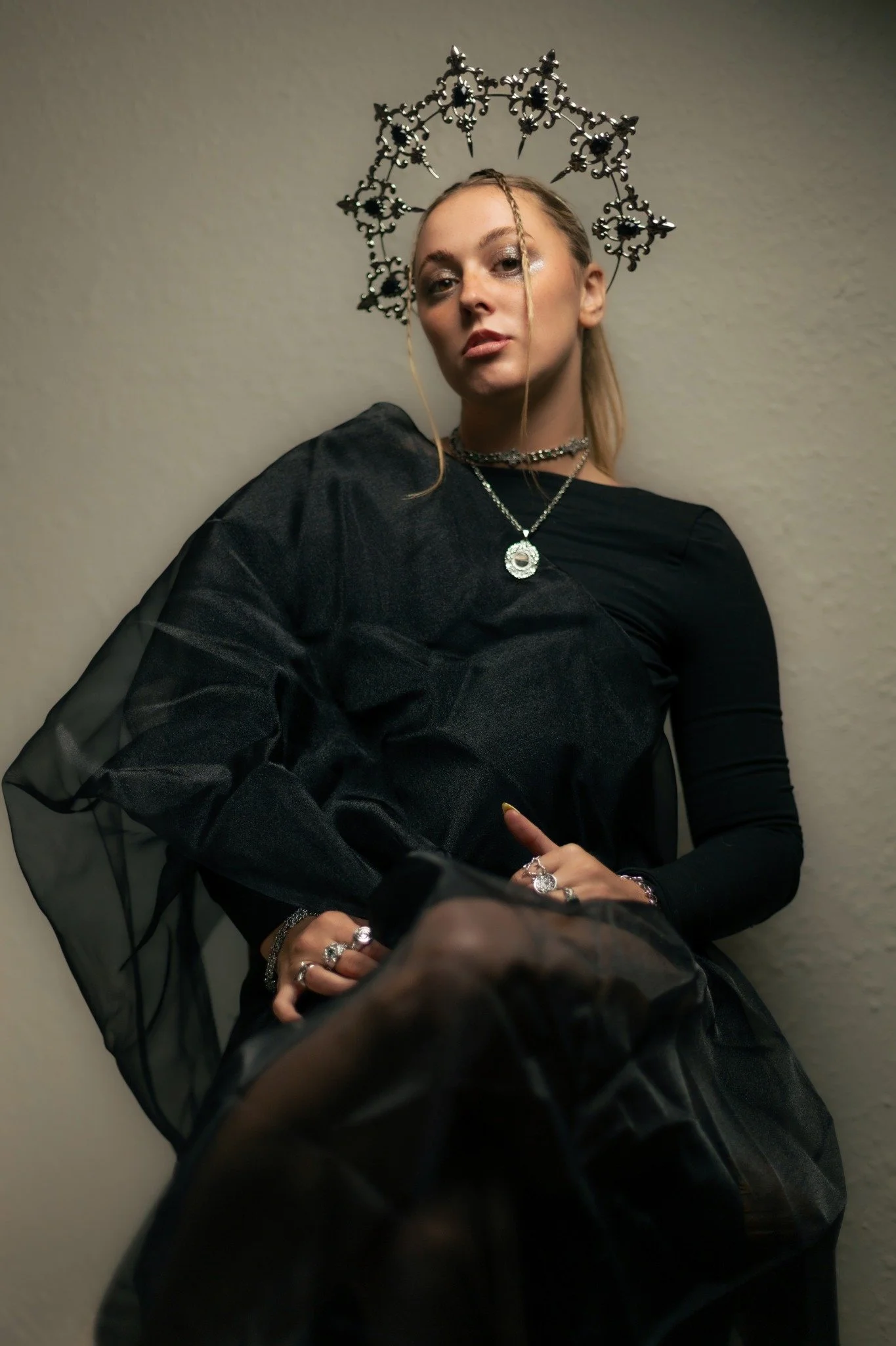 A woman with blonde hair, dressed in black, wearing silver jewelry and an ornate black crown, posing against a plain beige wall.
