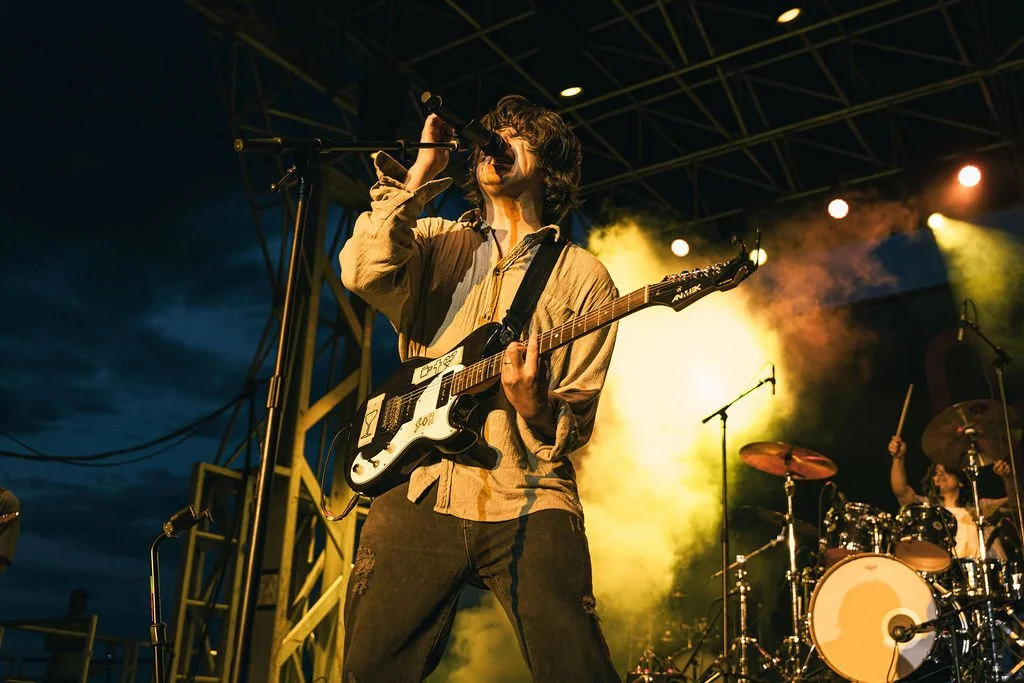A male musician singing into a microphone while playing an electric guitar on stage during a live outdoor concert at night, with colorful stage lighting and a drummer in the background.