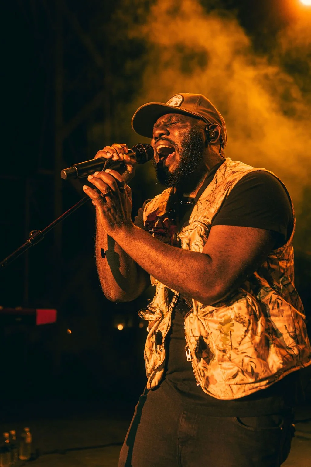 A male singer passionately performs on stage, holding a microphone with both hands, wearing a cap, a patterned vest, and black t-shirt, illuminated by warm orange stage lighting.
