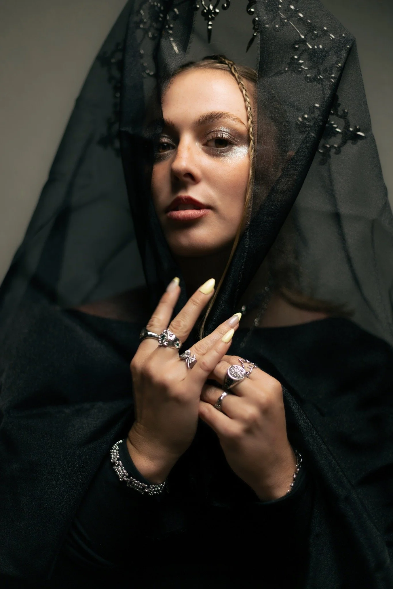 A woman partially obscured by black lace fabric with bead embellishments, wearing multiple rings, bracelets, and yellow nail polish, holding her hand near her face.