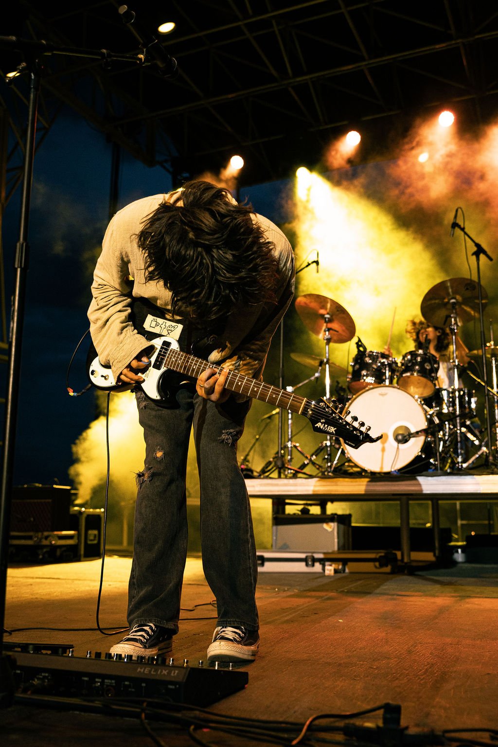 A musician with long black hair playing an electric guitar on stage, with a drummer behind them and colorful stage lighting.