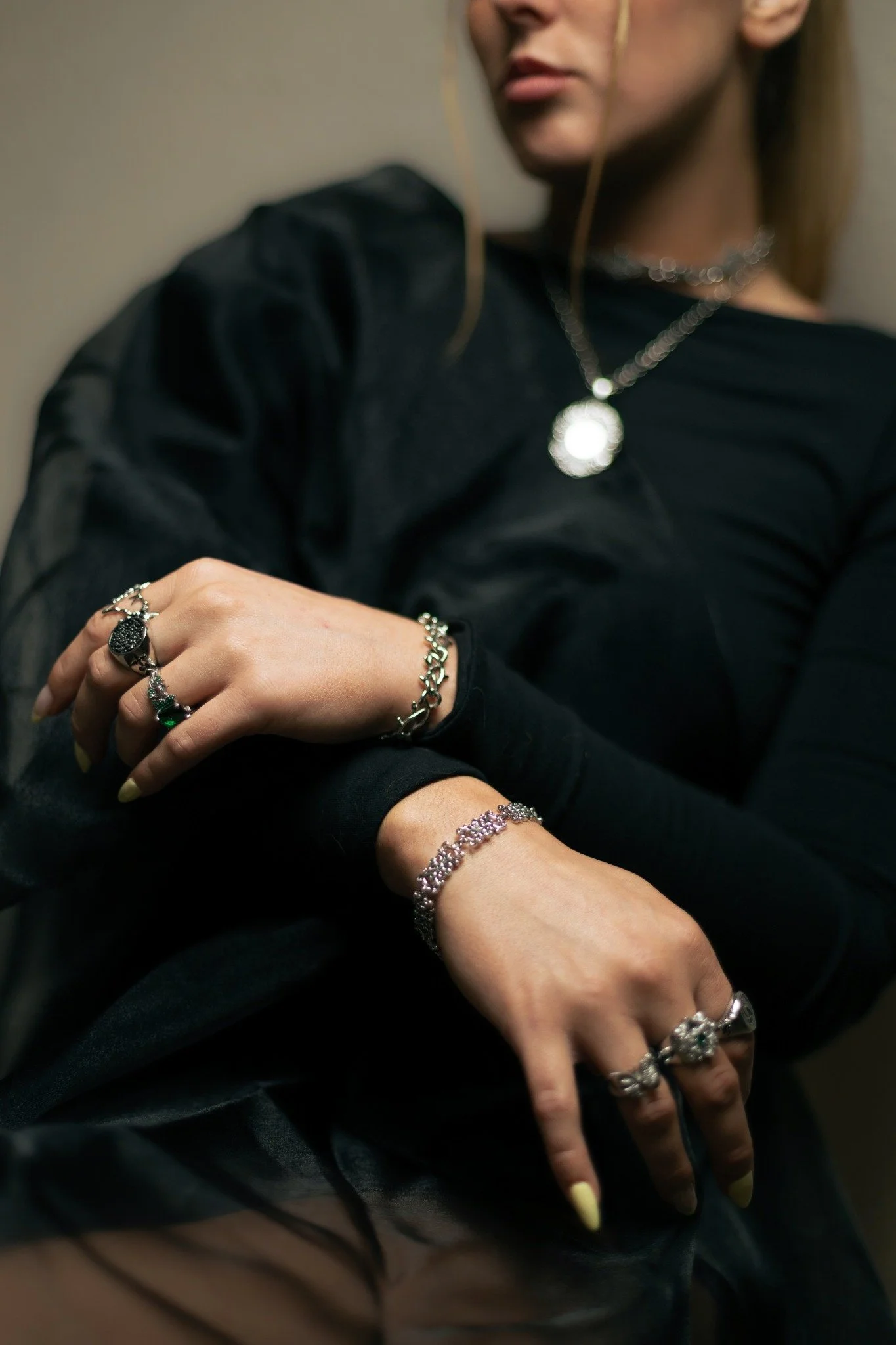 Close-up of a woman wearing multiple silver rings, bracelets, and a necklace, with her arms crossed and wearing a black sheer long sleeve shirt.
