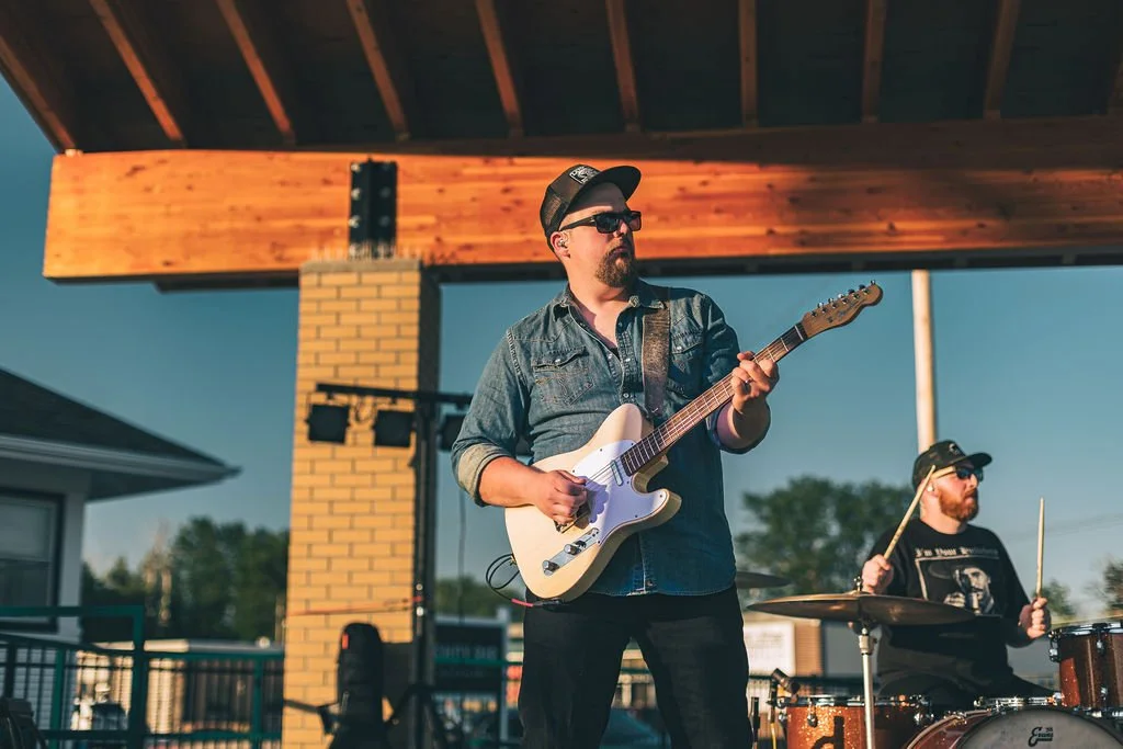 Two men playing music outdoors; one man is playing an electric guitar and the other is playing drums, both wearing sunglasses and casual clothing.