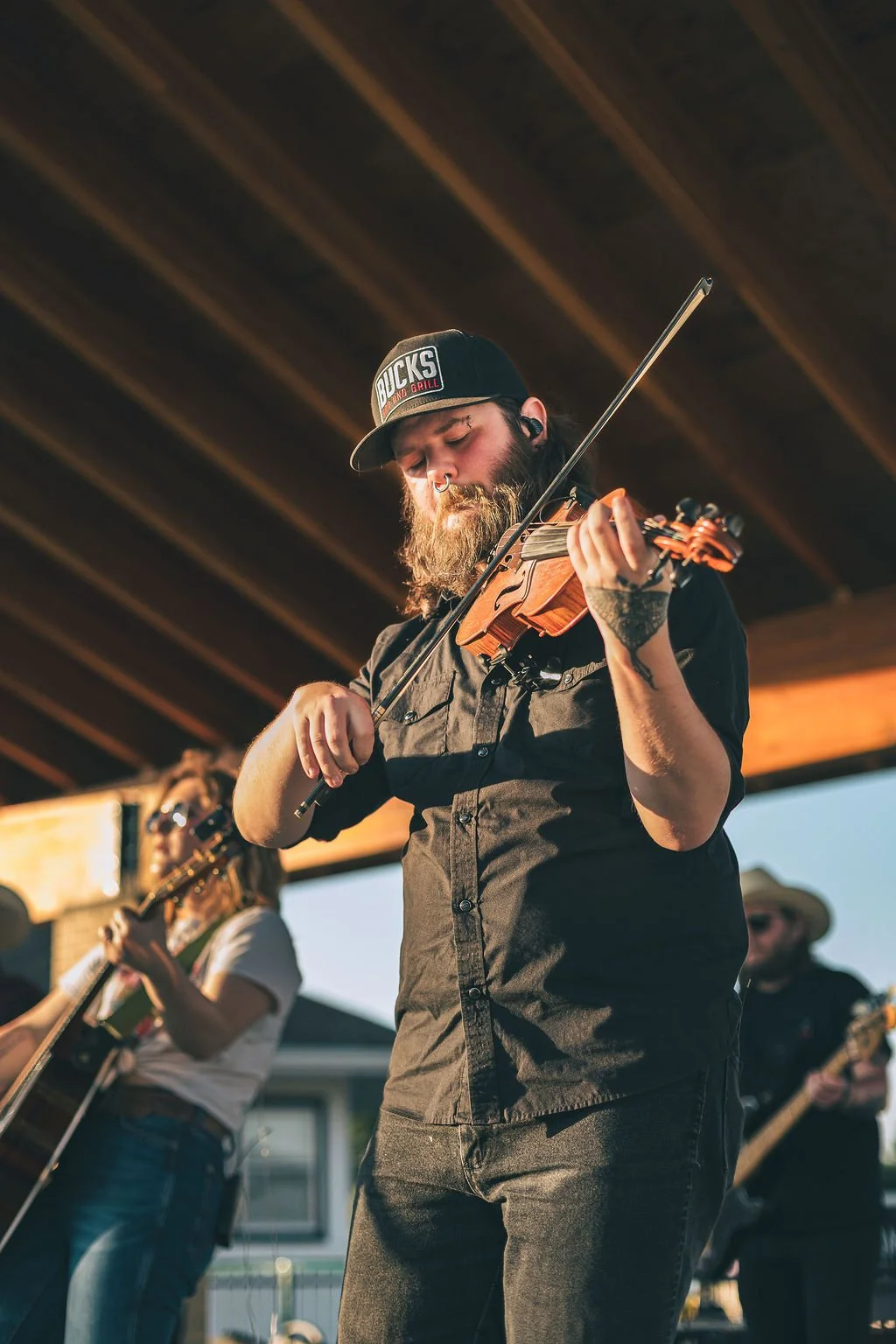 A man with a beard wearing a black cap with 'Bucks Bar and Grill' and playing a violin during an outdoor performance, with other musicians in the background under a wooden roof