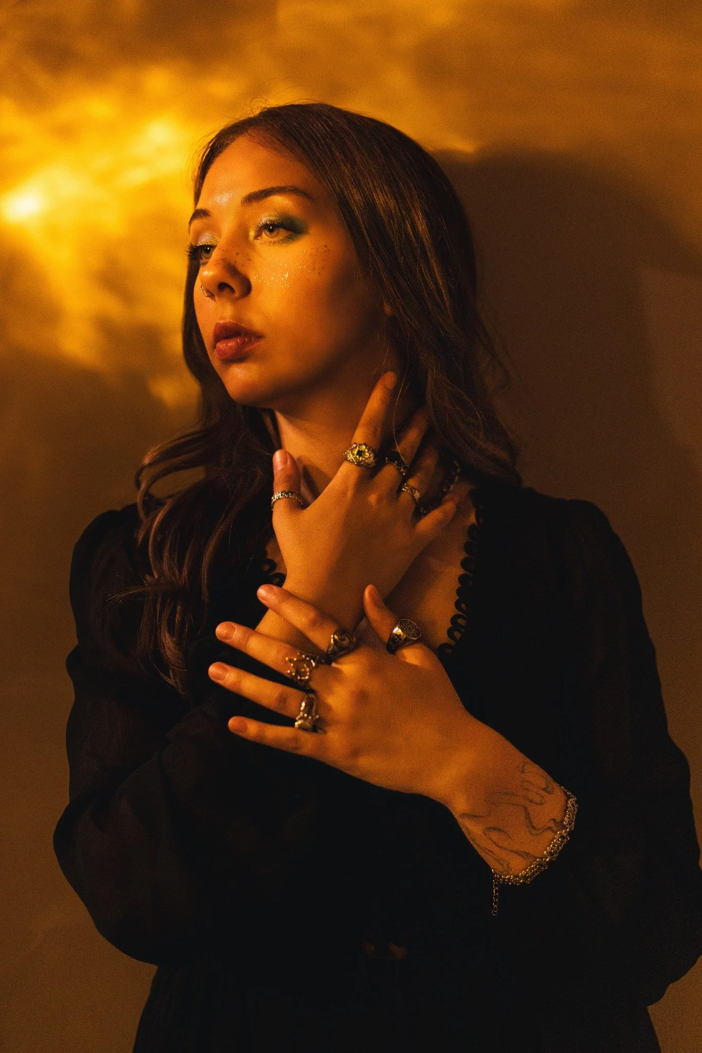 A woman with brown hair and blue eyes, wearing several rings and bracelets, has her hand near her face, with glitter on her cheek and a warm, golden background.