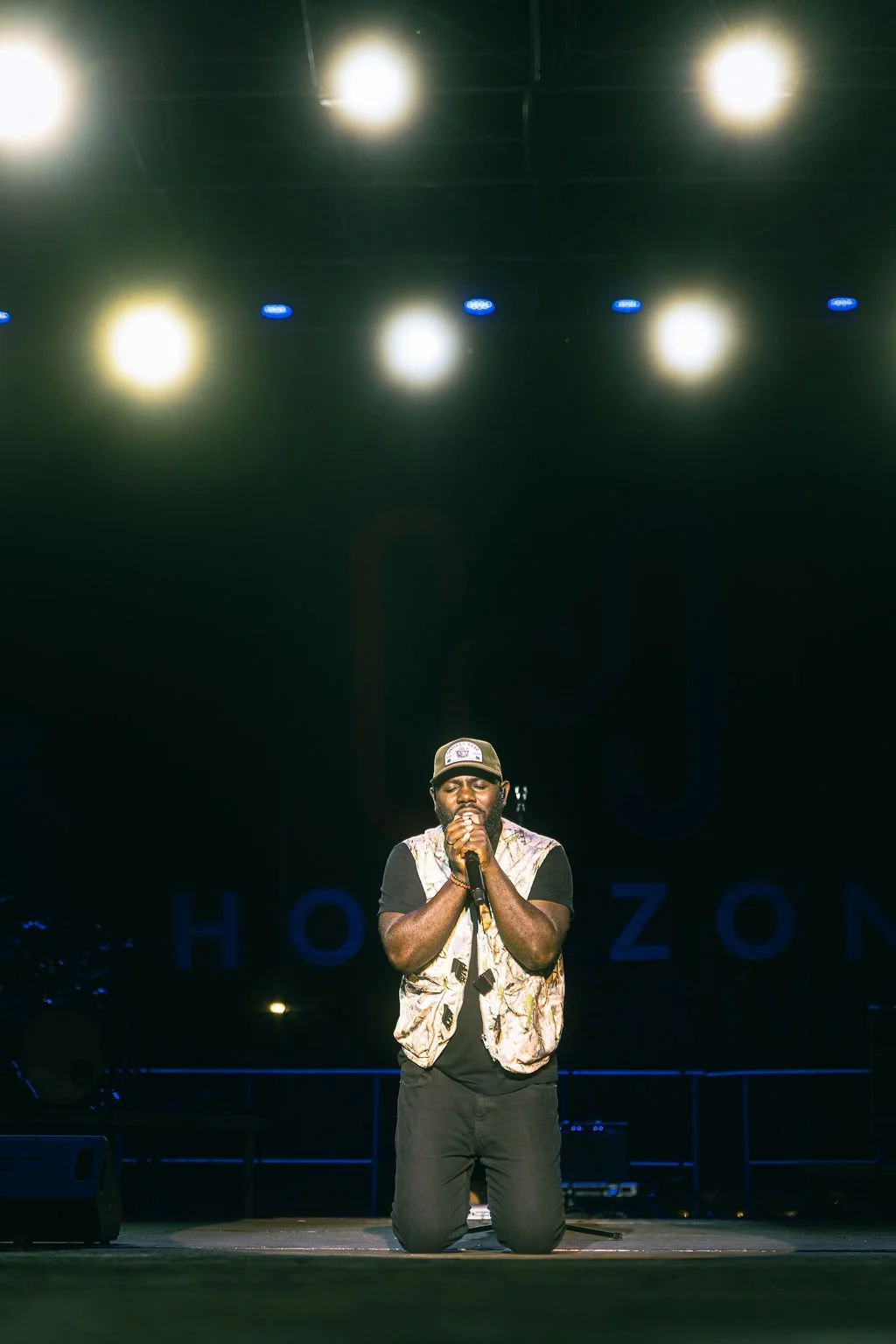 A male performer kneeling on stage, holding a microphone close to his mouth, with bright stage lights above him.