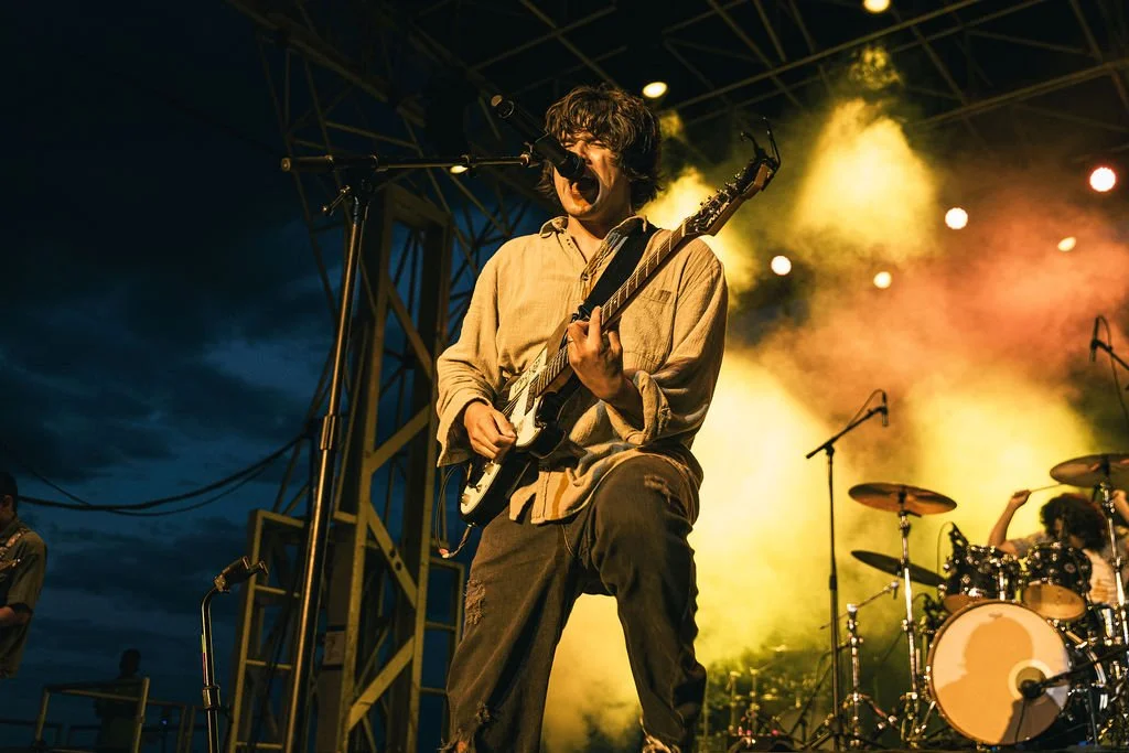 Musician singing into a microphone while playing an electric guitar on stage at a concert during sunset or nighttime with colorful stage lights.