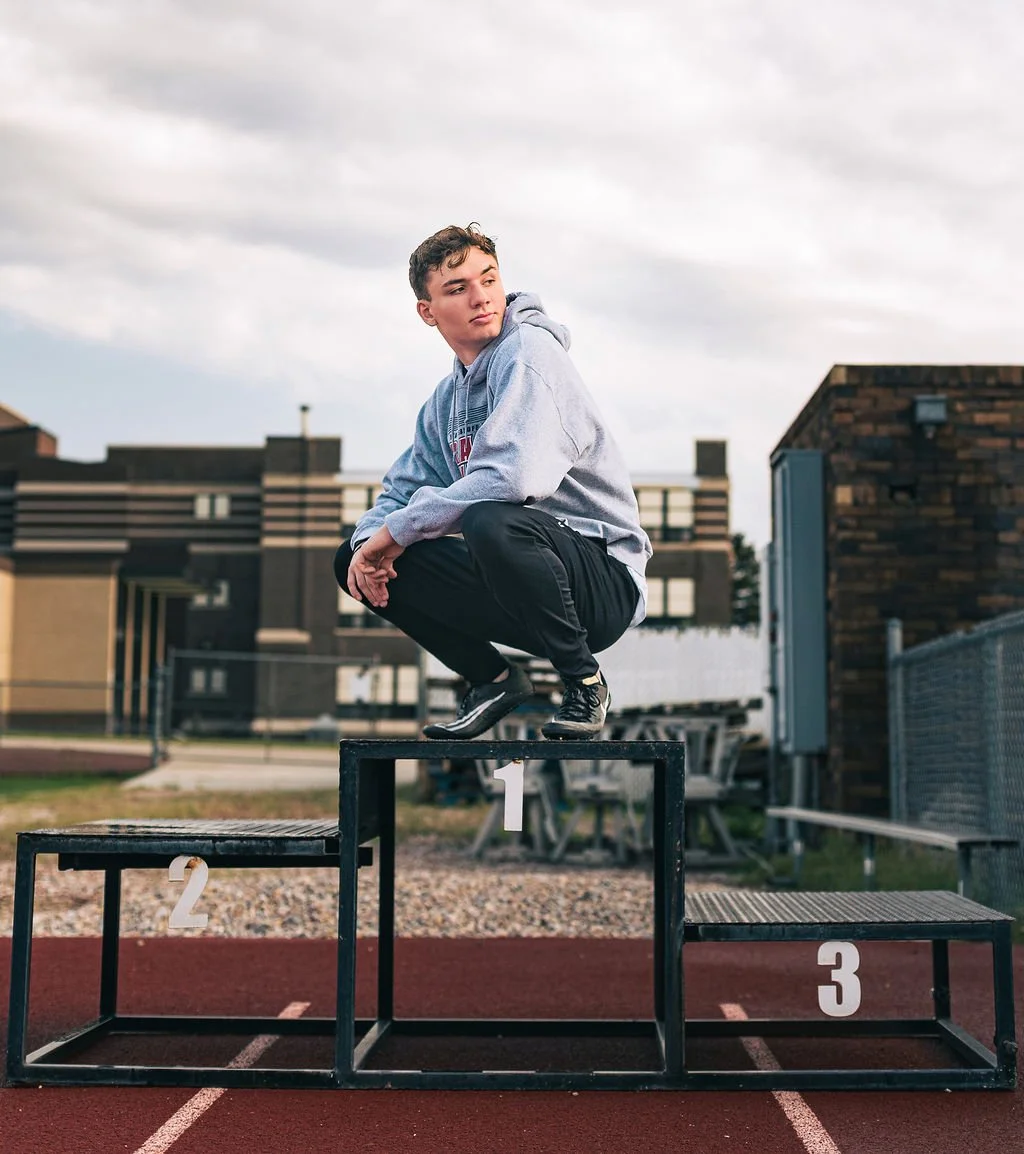 Young male athlete squatting on a first-place winners' podium at an outdoor track field, with buildings and cloudy sky in the background.