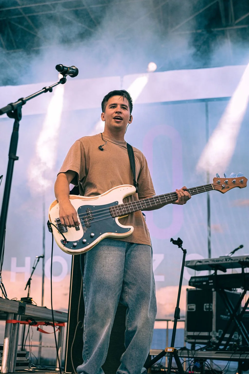 A young man playing bass guitar on stage during a live performance, with stage lights and musical equipment around him.