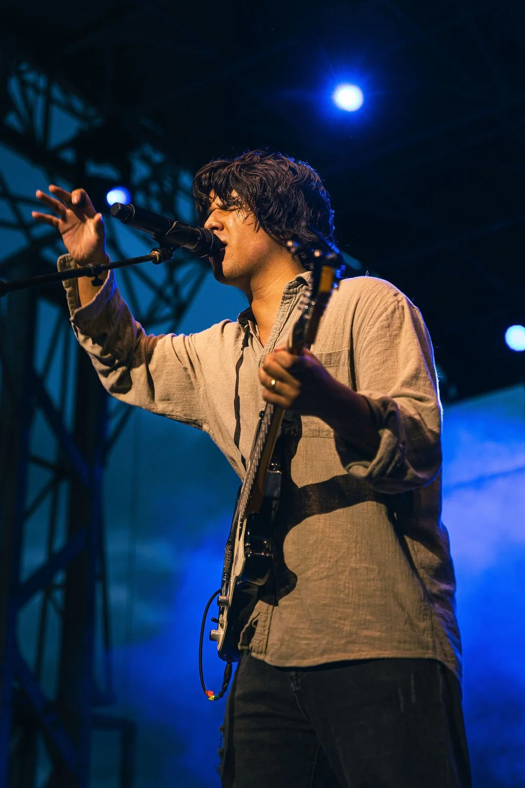 A male musician singing into a microphone and playing an electric guitar on a stage with blue lights