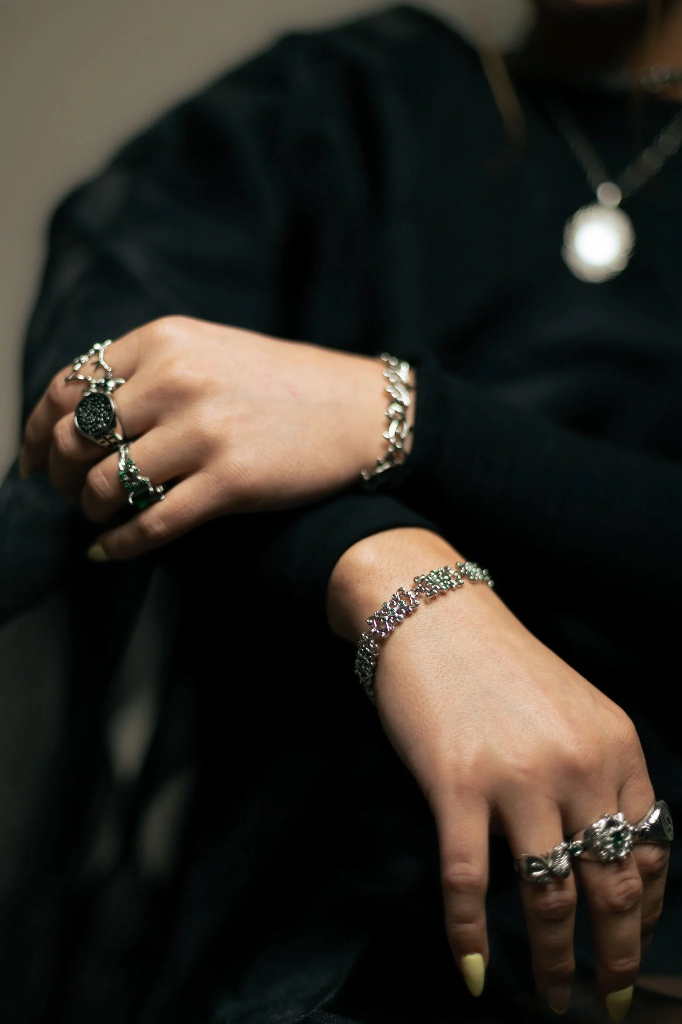 Close-up of a person wearing multiple silver jewelry rings, bracelets, and a necklace, with folded arms and crossed fingers, against a dark background.