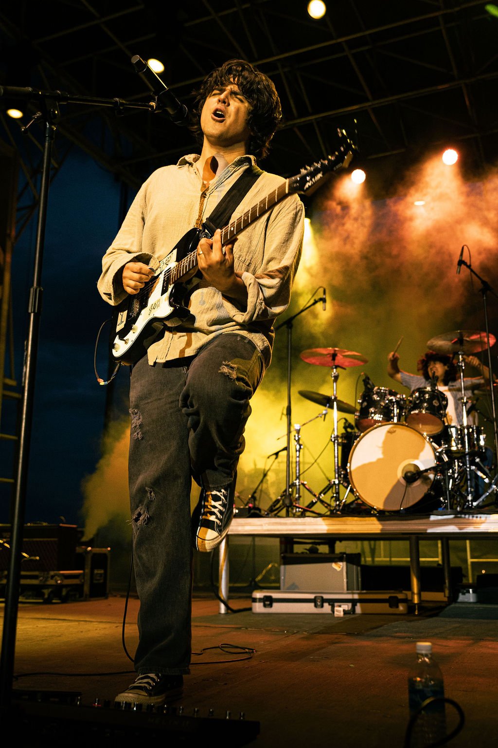 A young man with curly hair singing and playing an electric guitar on stage, with a drummer in the background and colorful stage lighting.