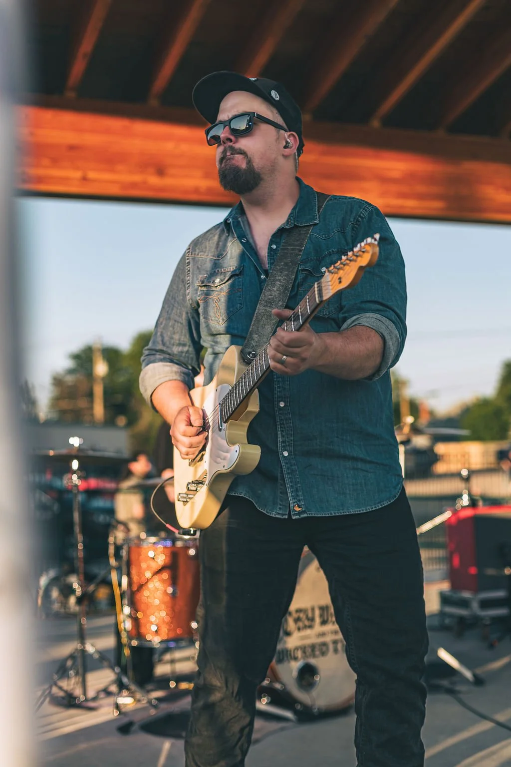 A man wearing sunglasses, a denim jacket, and a black cap playing an electric guitar during an outdoor performance with drums in the background.