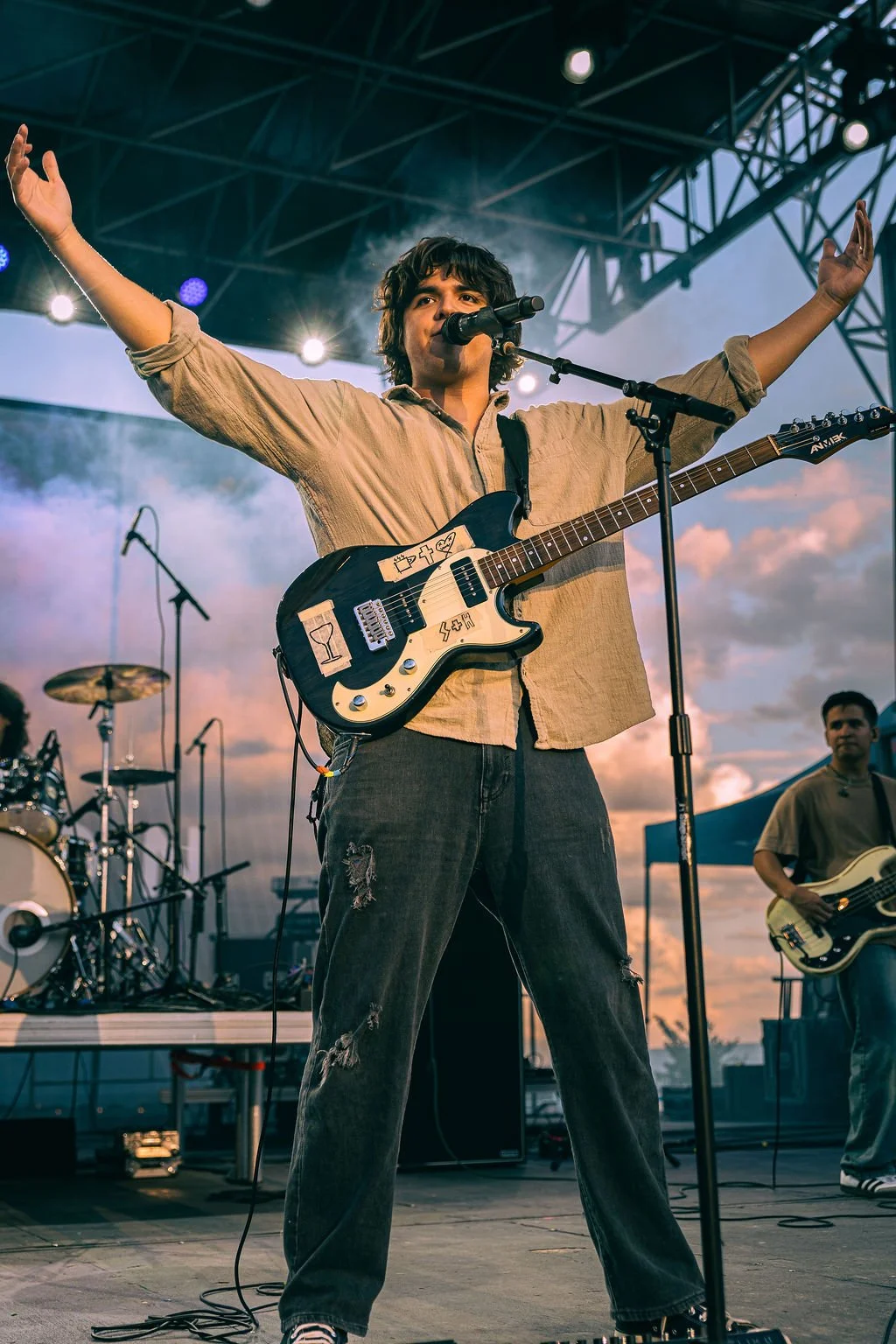A male musician with curly hair and a beige shirt performing on stage with a black and white electric guitar at sunset, with his arms raised and a drum set and another band member visible in the background.
