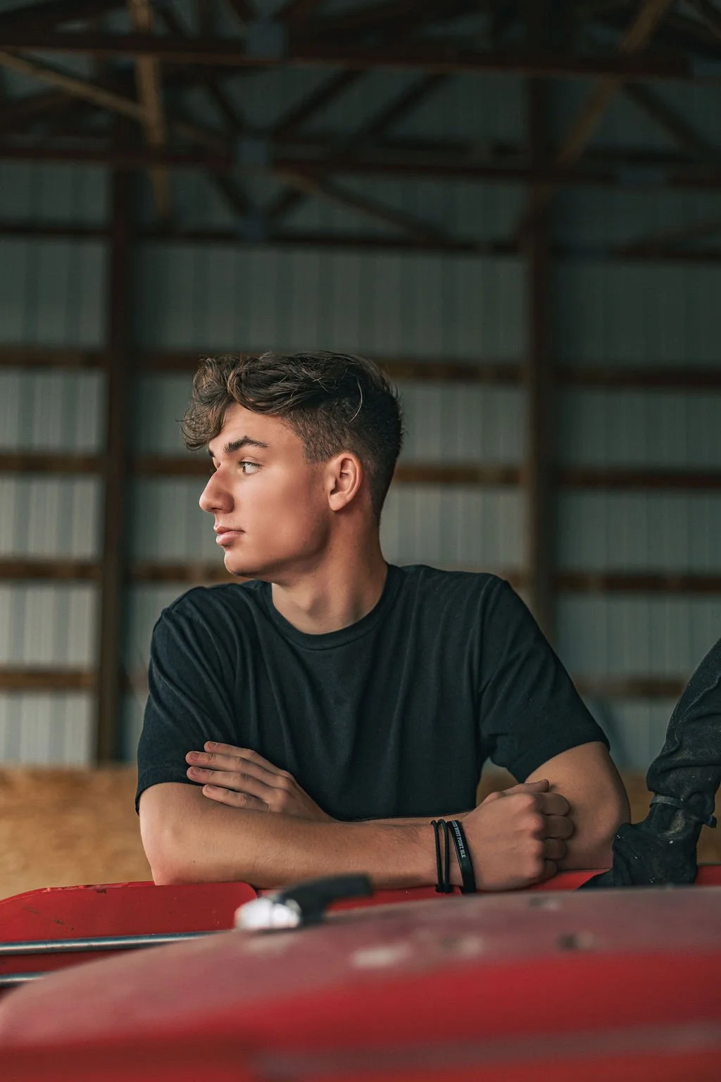 A young man with short curly hair, wearing a black t-shirt and black wristbands, sits with arms crossed in a barn or warehouse with metal and wood walls.