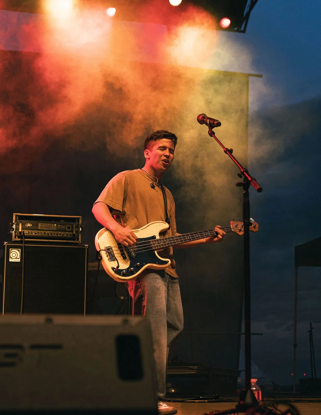 A young man plays a bass guitar on stage during a concert, with colorful stage lights and equipment in the background.