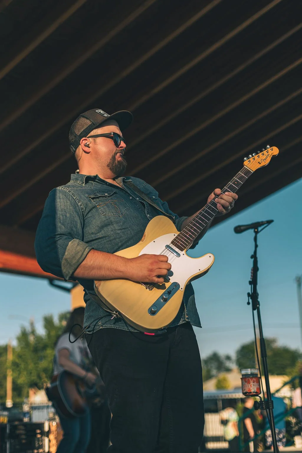 A man with sunglasses, a baseball cap, and a denim shirt playing an electric guitar at an outdoor concert.