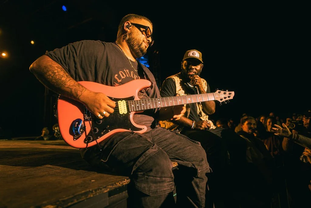 Two musicians performing on stage, one playing an electric guitar and the other singing into a microphone, with an audience watching.