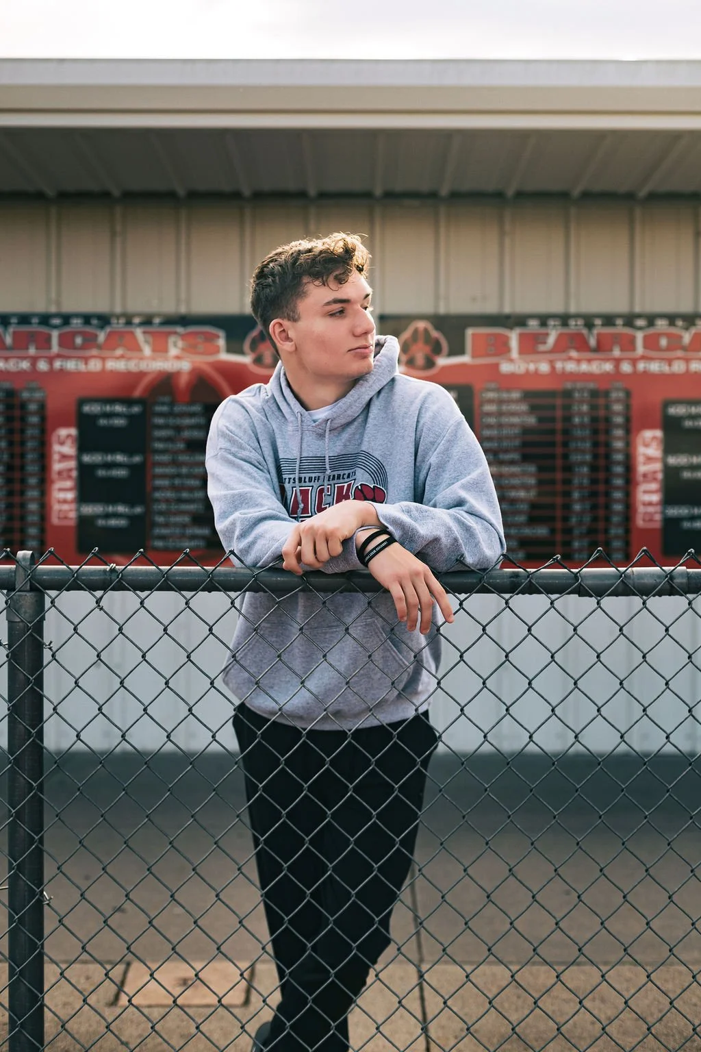 A young man with curly hair wearing a gray hoodie and black pants leaning on a chain-link fence at a sports field, with scoreboard in the background.