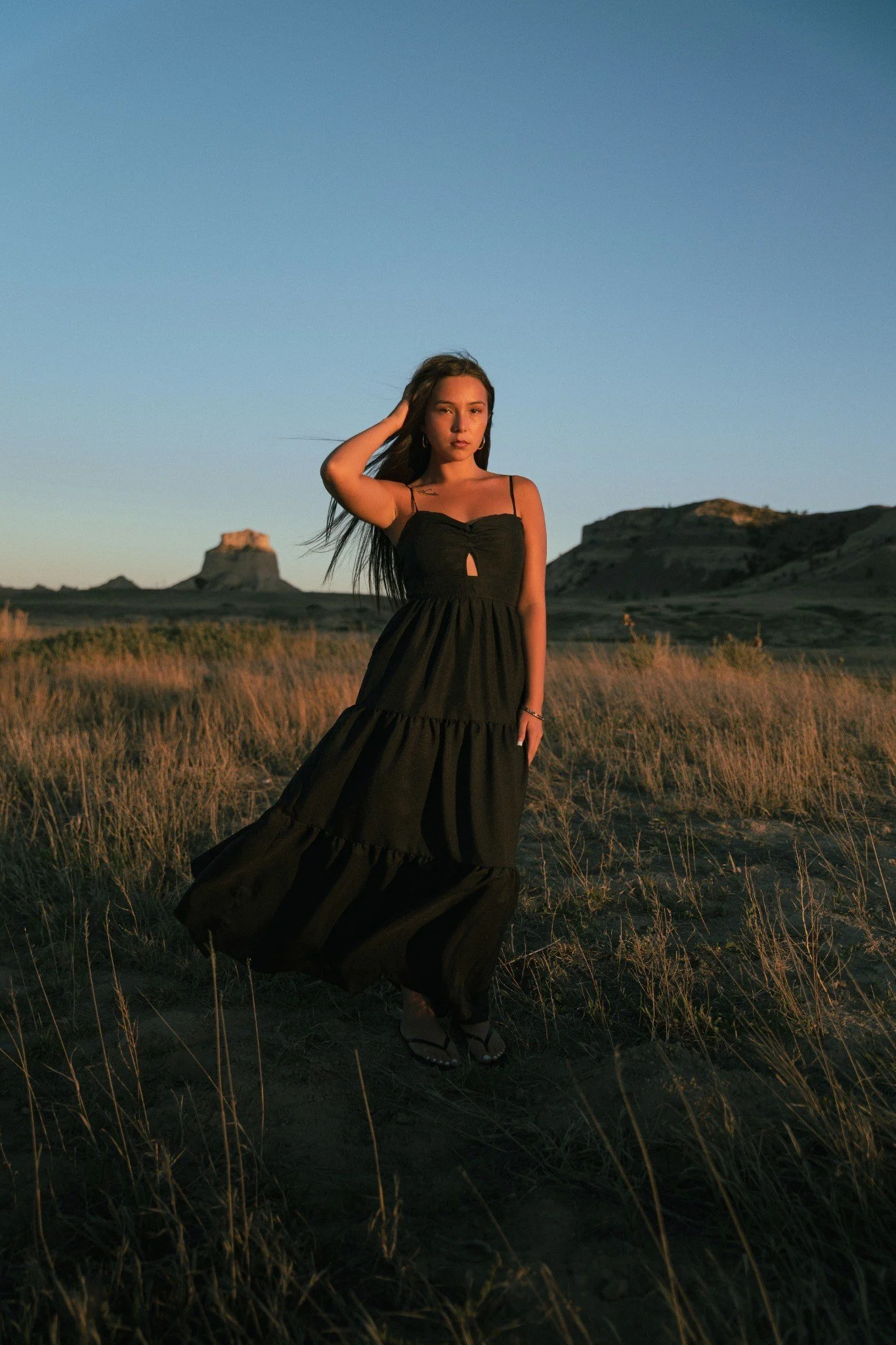 A woman standing in a grassy field with highland formations in the background at sunset, wearing a long black dress and sandals.