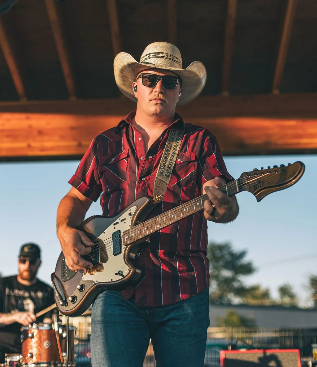 A man wearing a cowboy hat, sunglasses, and a red plaid shirt playing an electric guitar during an outdoor performance, with a drummer visible in the background.