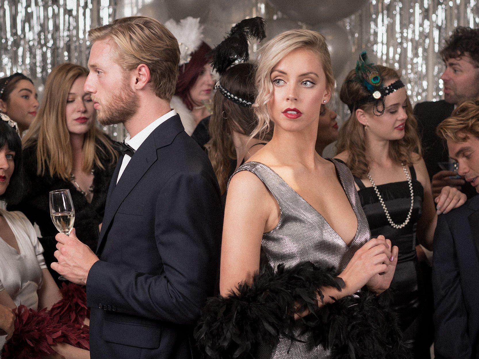 A woman in a silver dress with feathers at her waist stands among people dressed in vintage style at a fancy party. She has blonde hair styled in waves, bright red lipstick, and appears to be looking away. The man beside her in a tuxedo holds a champagne glass, with many other party guests in the background.