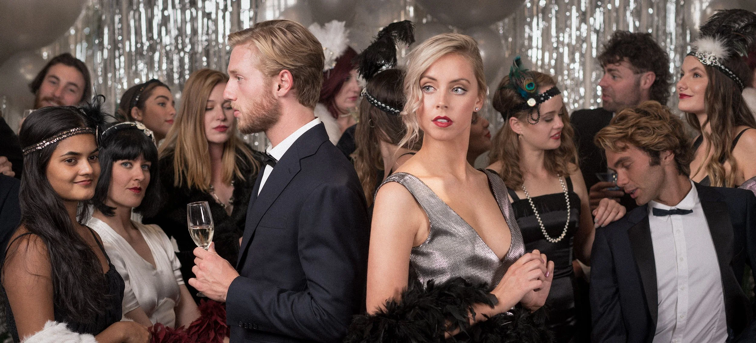 People attending a formal party or event, dressed in elegant evening wear, with some wearing feathered headbands, in a room decorated with silver streamers and balloons.