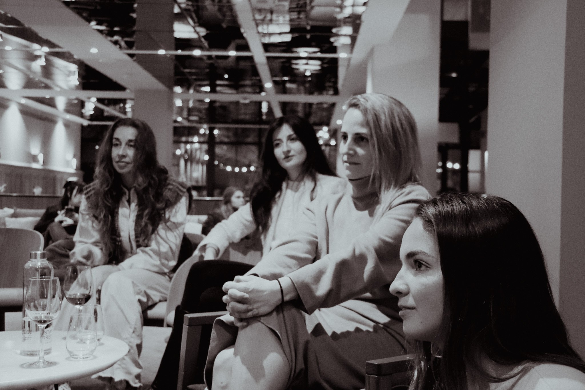 Group of four women sitting and conversing in a modern indoor setting, with drinks on a table in front of them.