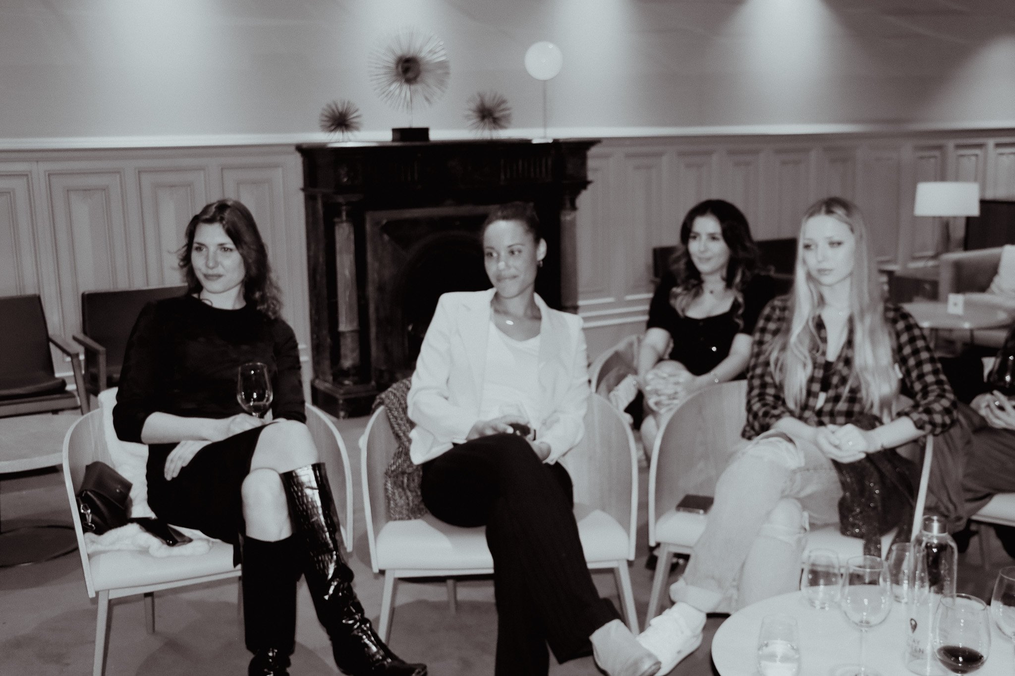 Four women sitting in chairs in a dimly lit room, with a decorative fireplace and modern decor in the background. One woman is holding a glass. The atmosphere appears relaxed and social.