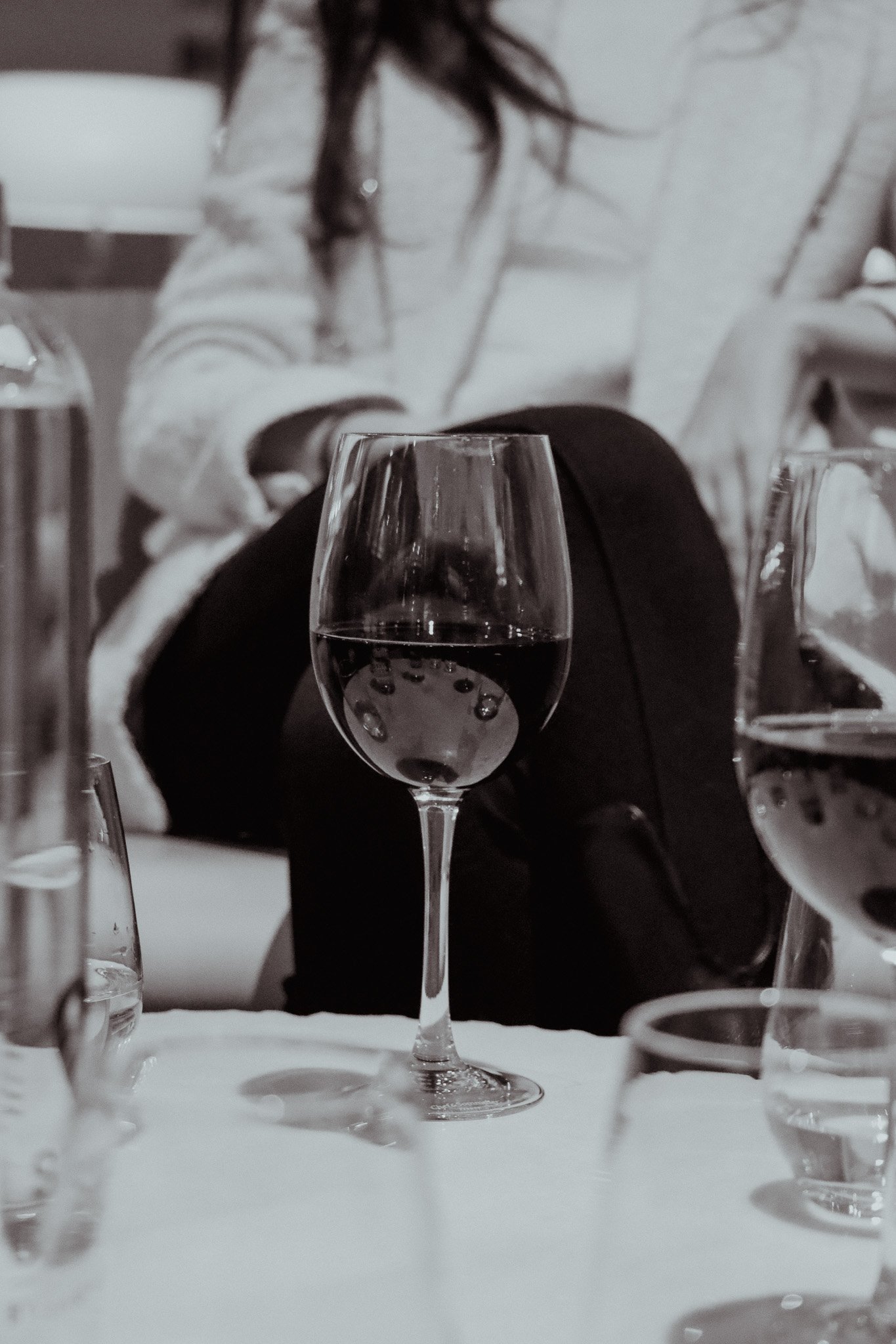 A black and white photograph of a wine glass on a table, with a seated person in the background and other glasses visible.