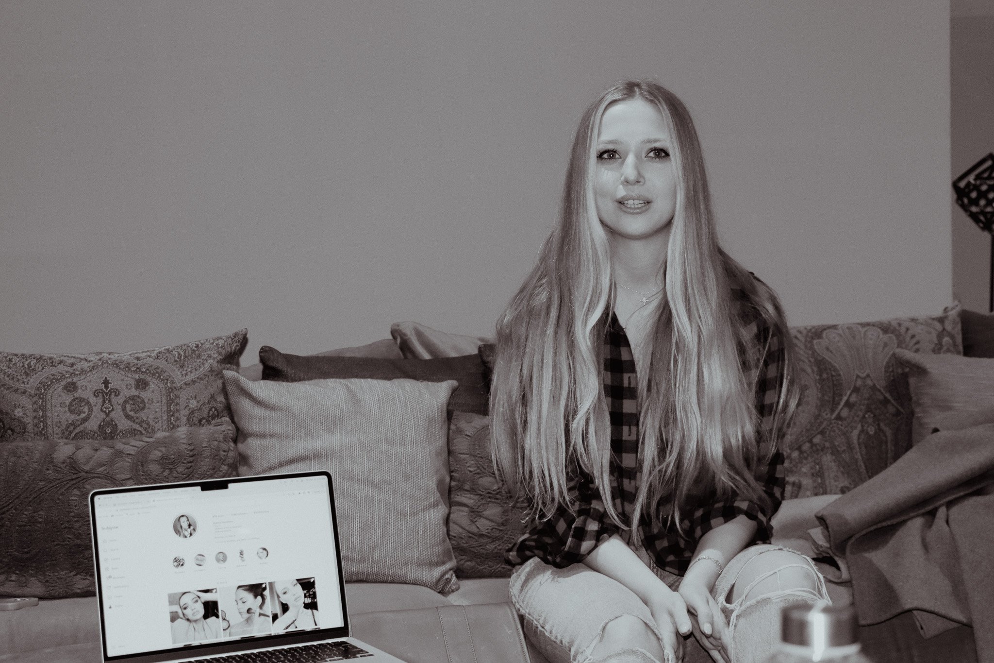 Black and white photo of a woman with long hair sitting on a sofa, laptop with Instagram profile visible, decorative pillows in the background.