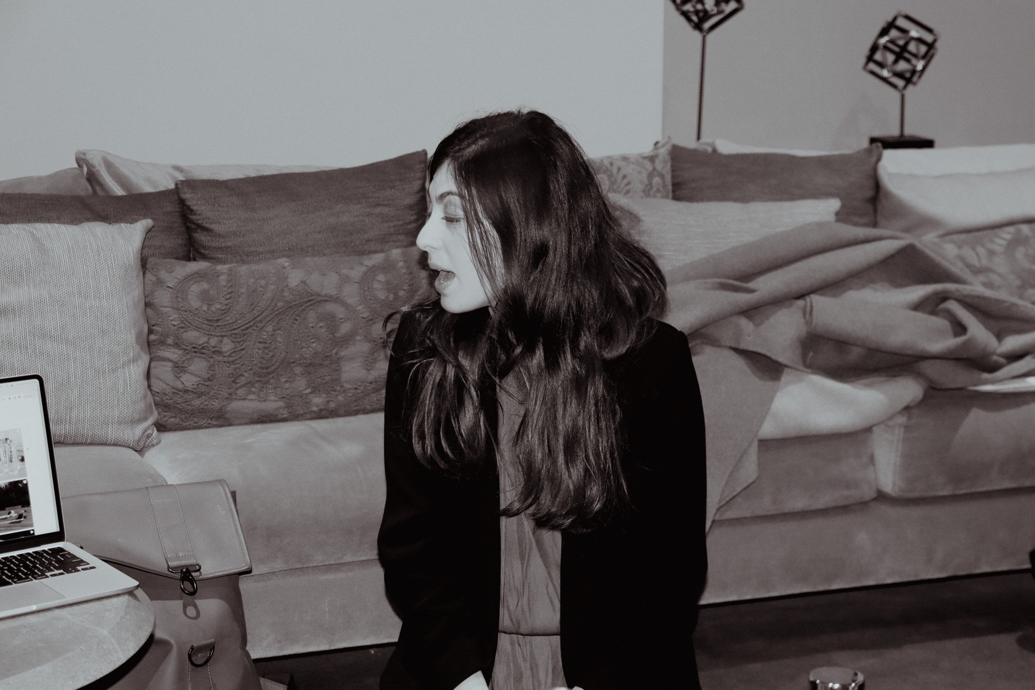 Black and white image of a woman with long hair sitting on a sofa, turned in profile. A laptop and bag are on a table beside her. Decorative pillows are on the sofa.