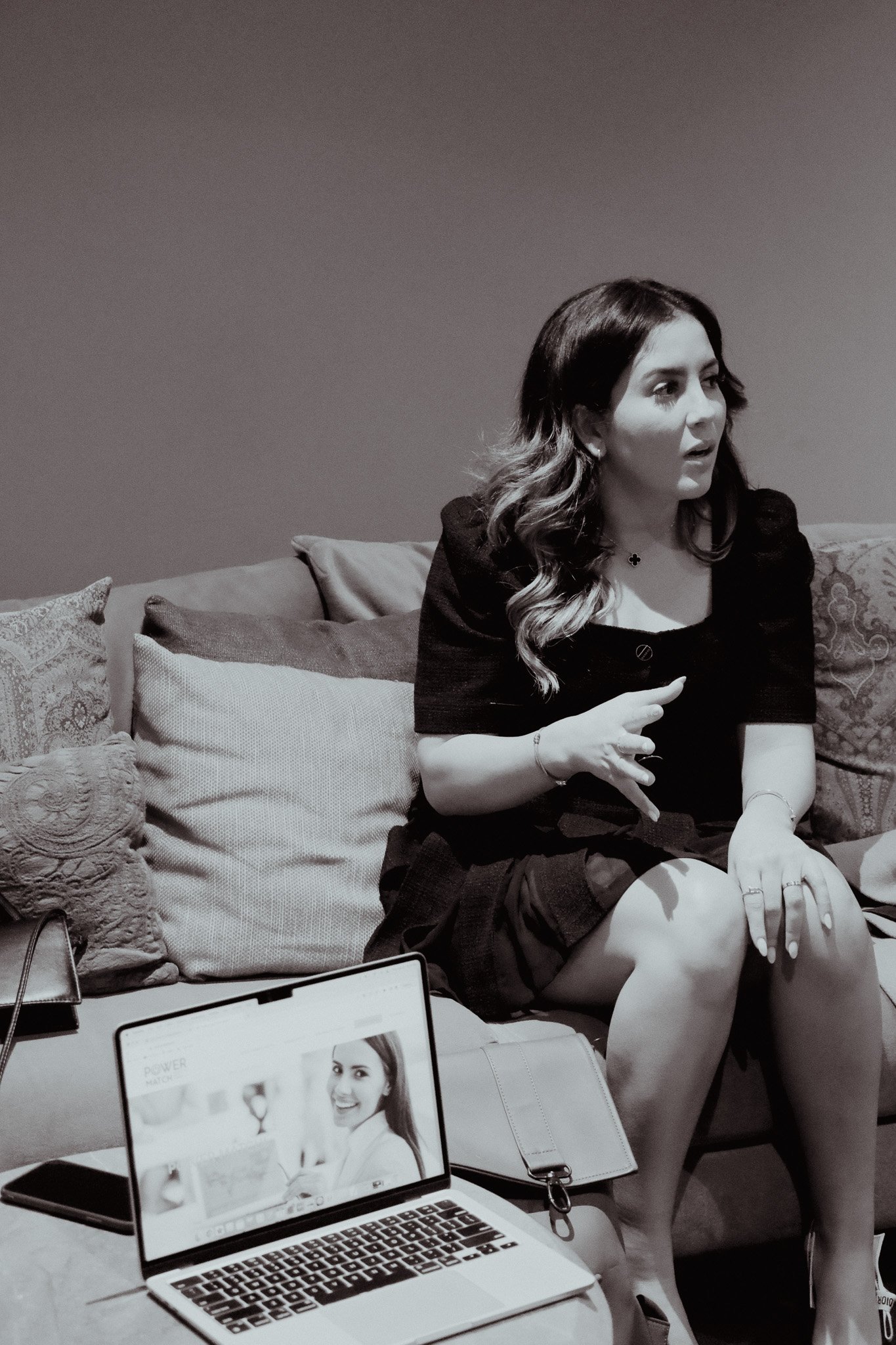 Woman sitting on a couch, talking with gestures, next to an open laptop displaying a smiling woman's image.
