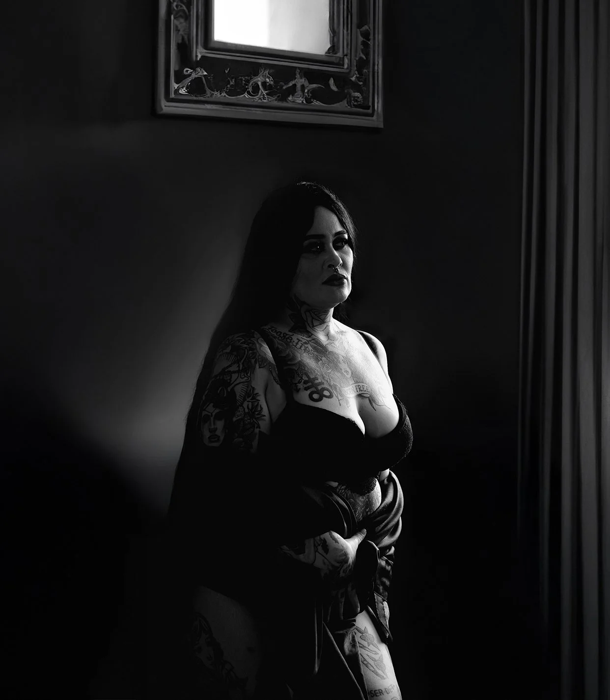 A woman with tattoos standing in a dimly lit room, partly illuminated by natural light from a window.