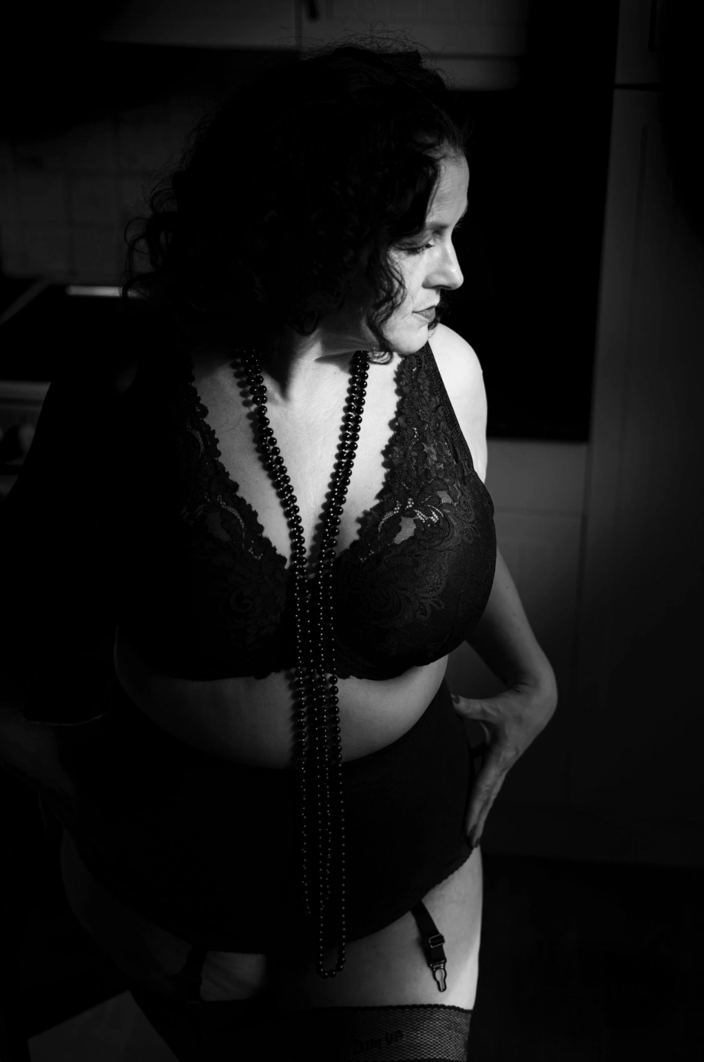 A black and white photo of a woman with dark curly hair, wearing lace lingerie and pearls, standing in a room with one hand on her hip and looking down.