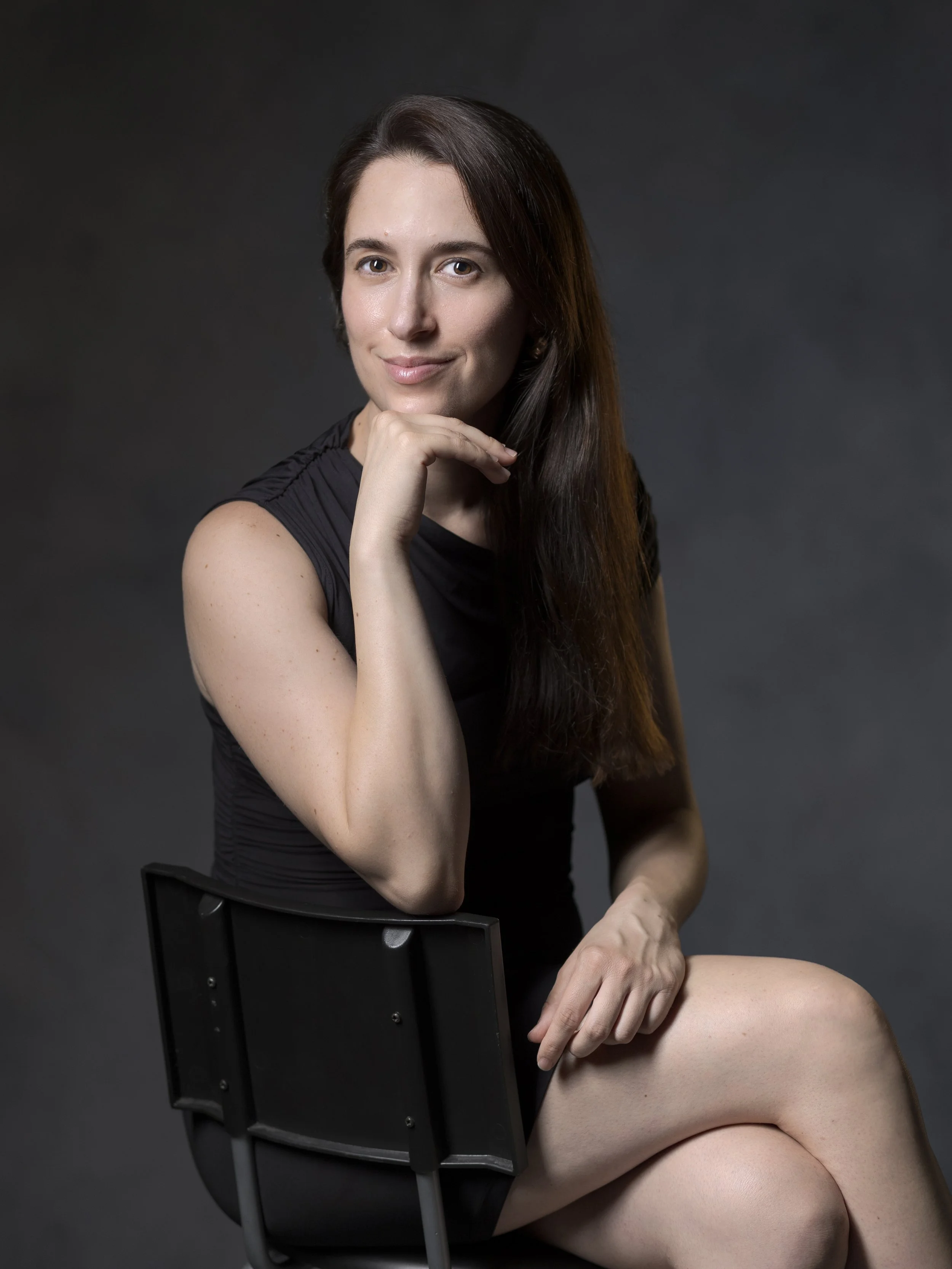 Retrato de una mujer con cabello largo, posando con la mano en la barbilla, sentada en una silla negra, vestida con camiseta negra, con un fondo oscuro.