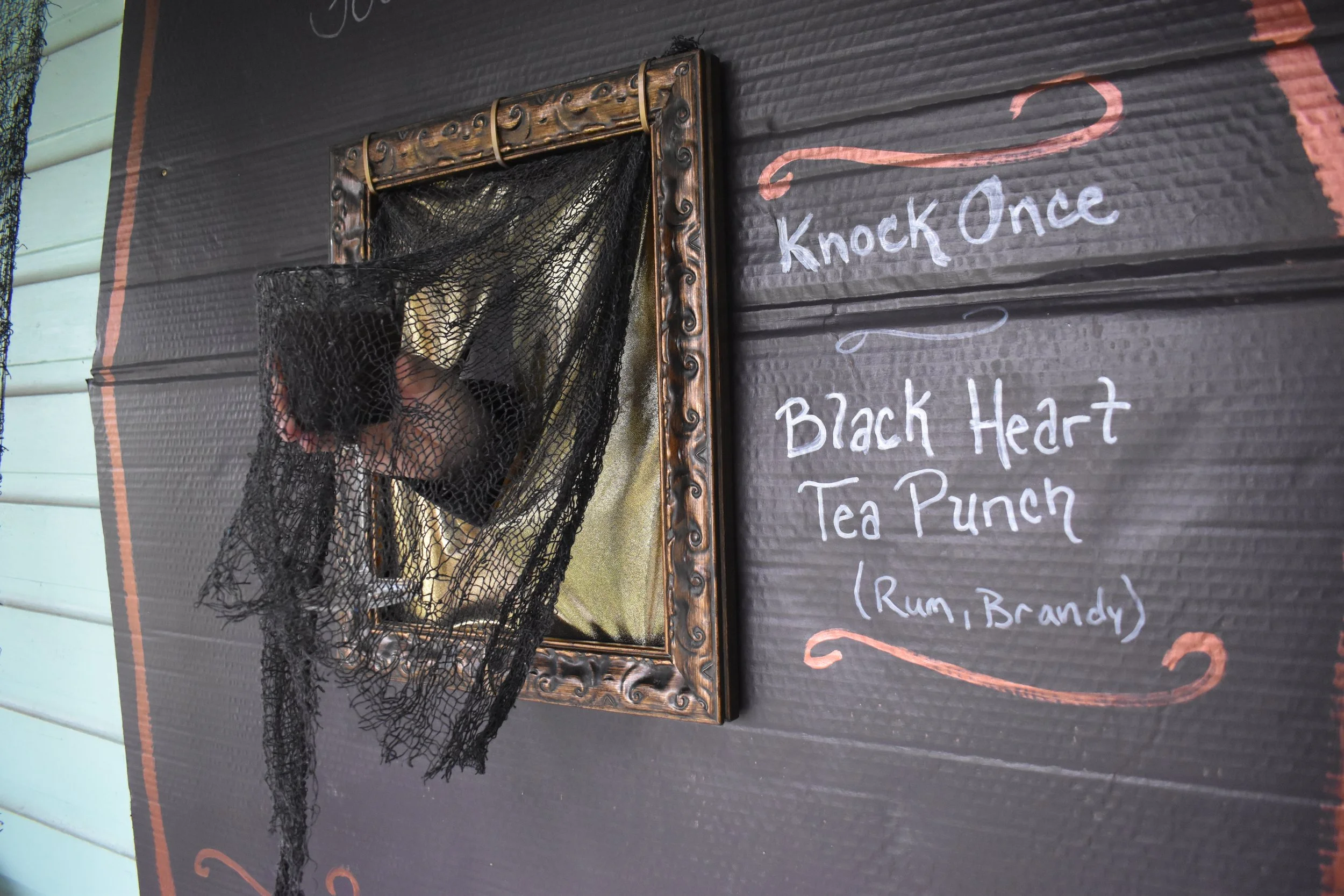 close-up photo of carboard coffin lid with a hand holding a drink stretching out through a framed hole.