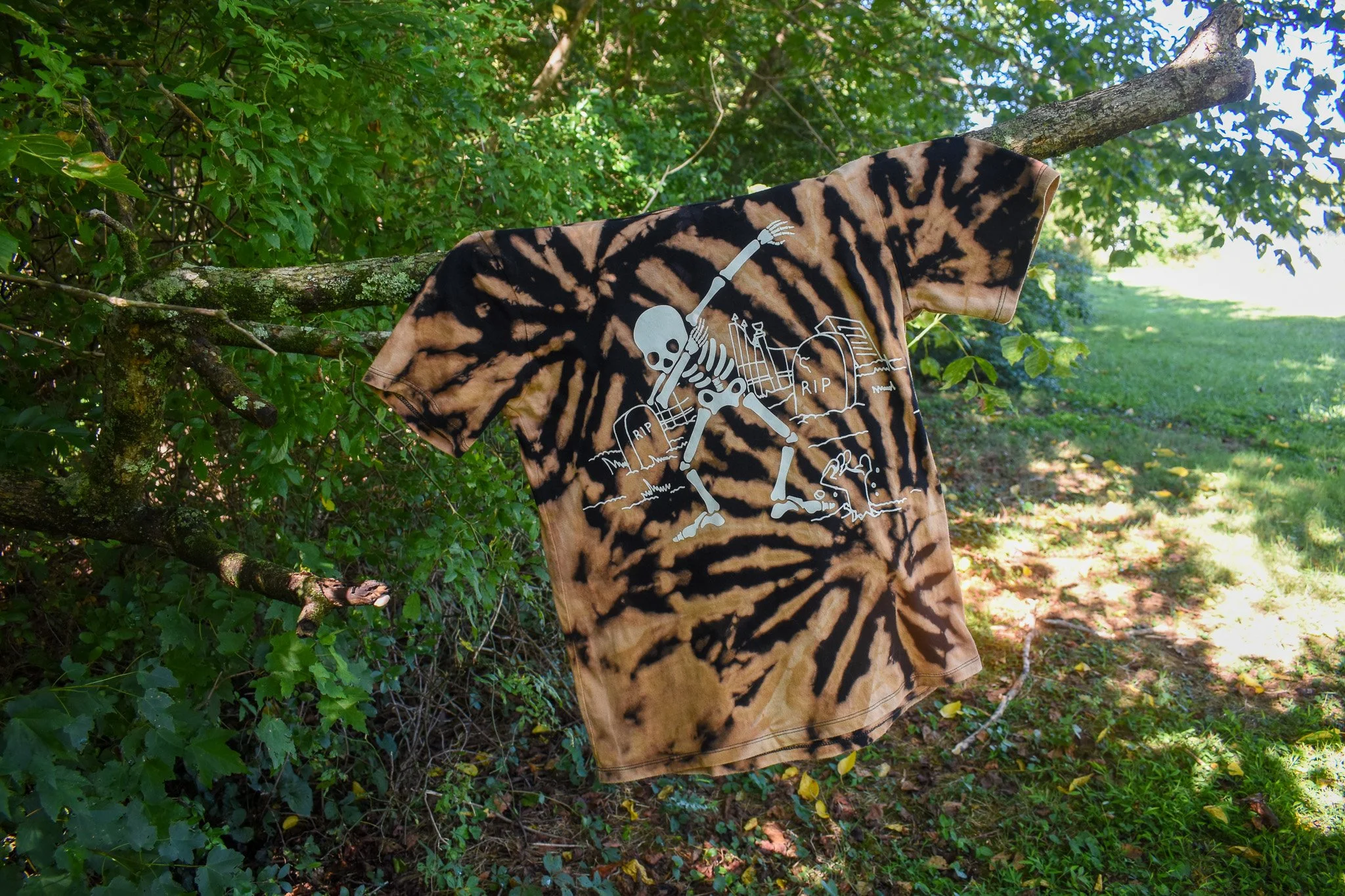 A bleach dyed t-shirt with a dabbing skeleton image hangs on a tree branch