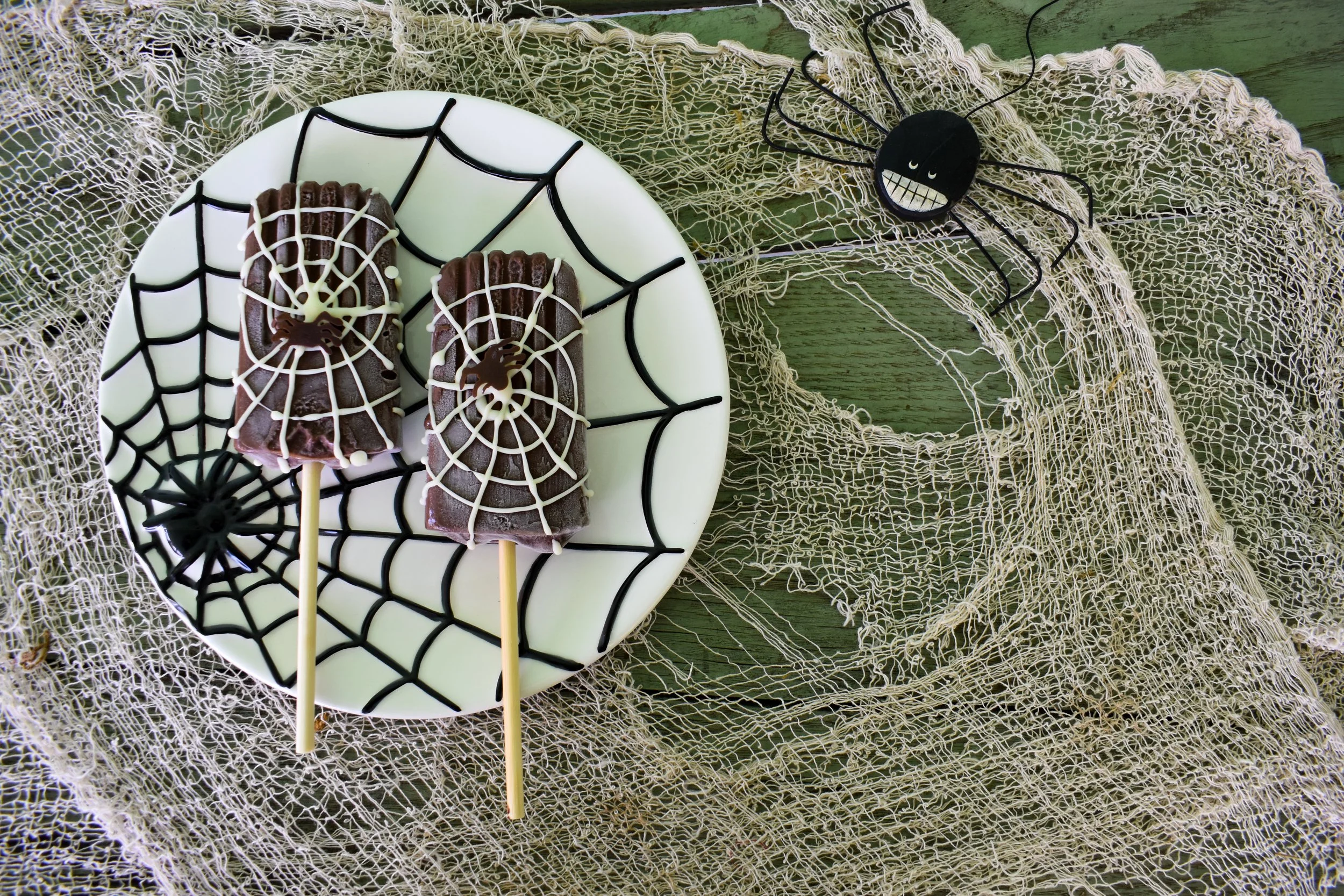 Two popsicles decorated with white chocolate spider webs rest on a spiderweb plate.