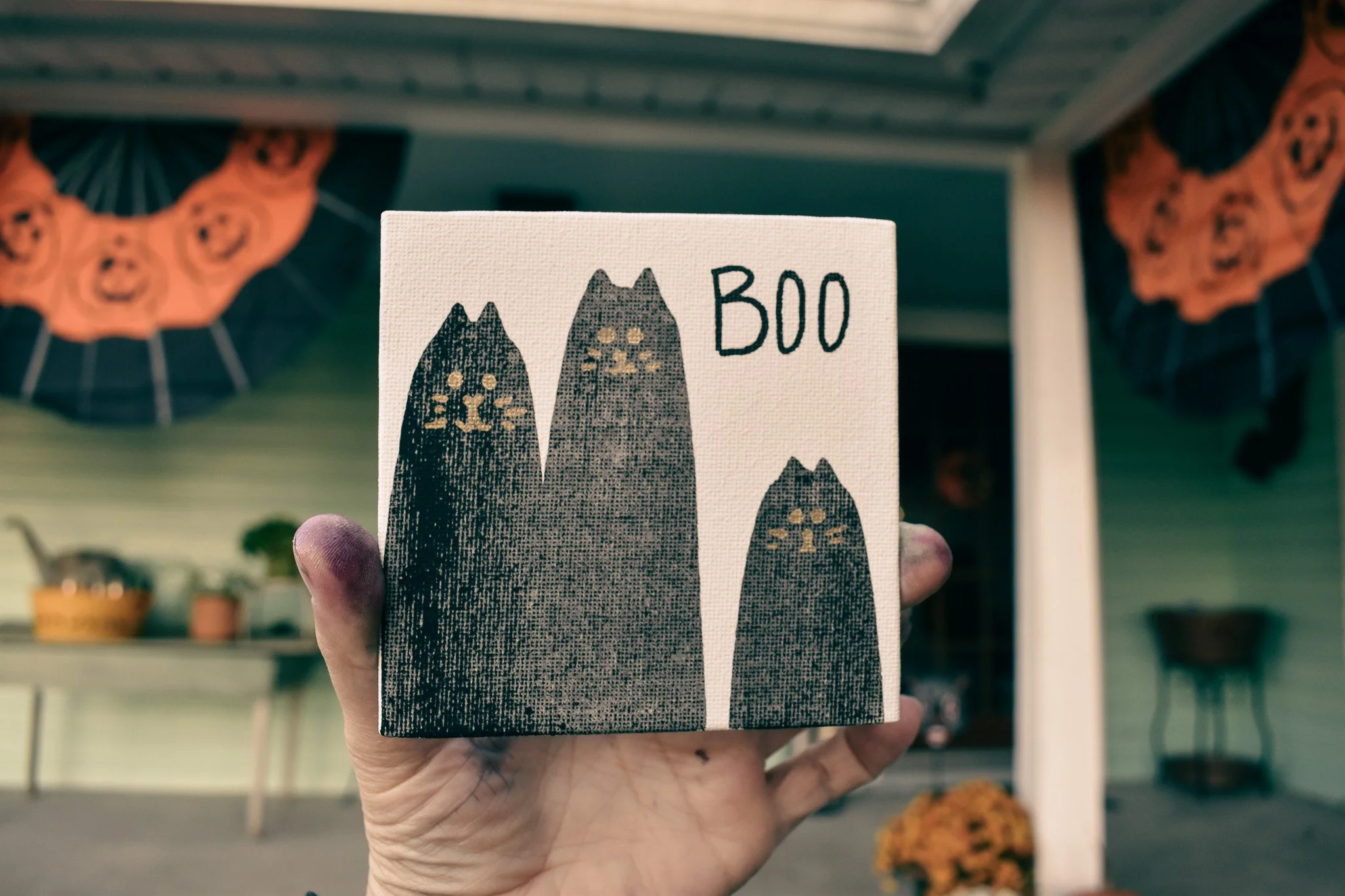 A hand holds up a mini canvas with three black cat shapes and the word "boo"