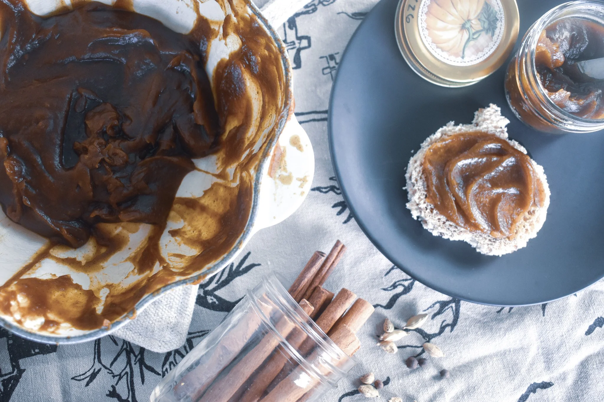Overhead view a splatter covered pot of pumpkin butter, a plate with a pumpkin shaped toast with pumpkin butter, and spices spilling onto the table