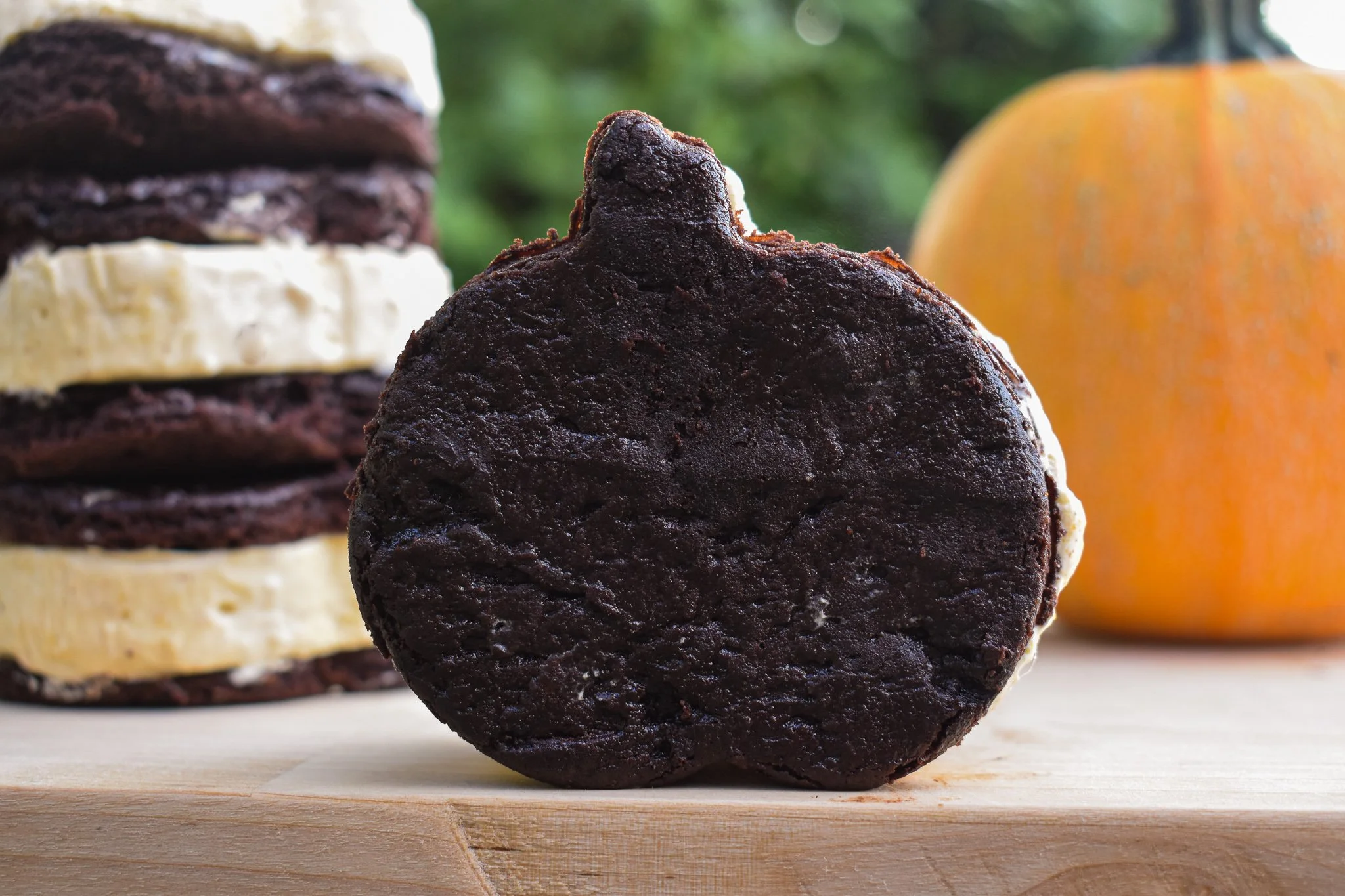 pumpkin shaped ice cream sandwich on a wooden tray with a pumpkin and sandwich stack behind it