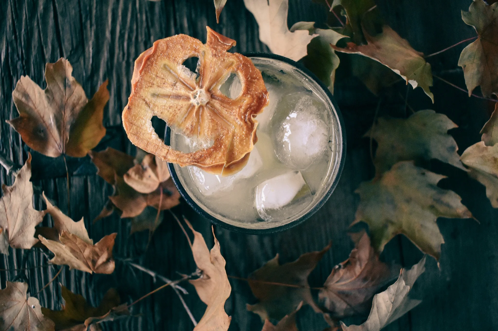 glass sits on a wooden table covered in autumn leaves; glass is garnished with a wrinkly jack-o-lantern face made of dried persimmon.