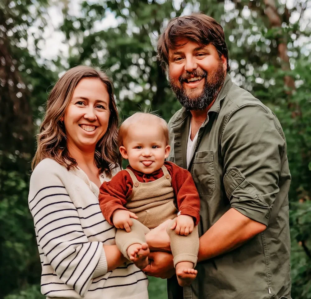A happy family of three outdoors in a green, wooded area, with the mother, father, and baby holding the little boy.