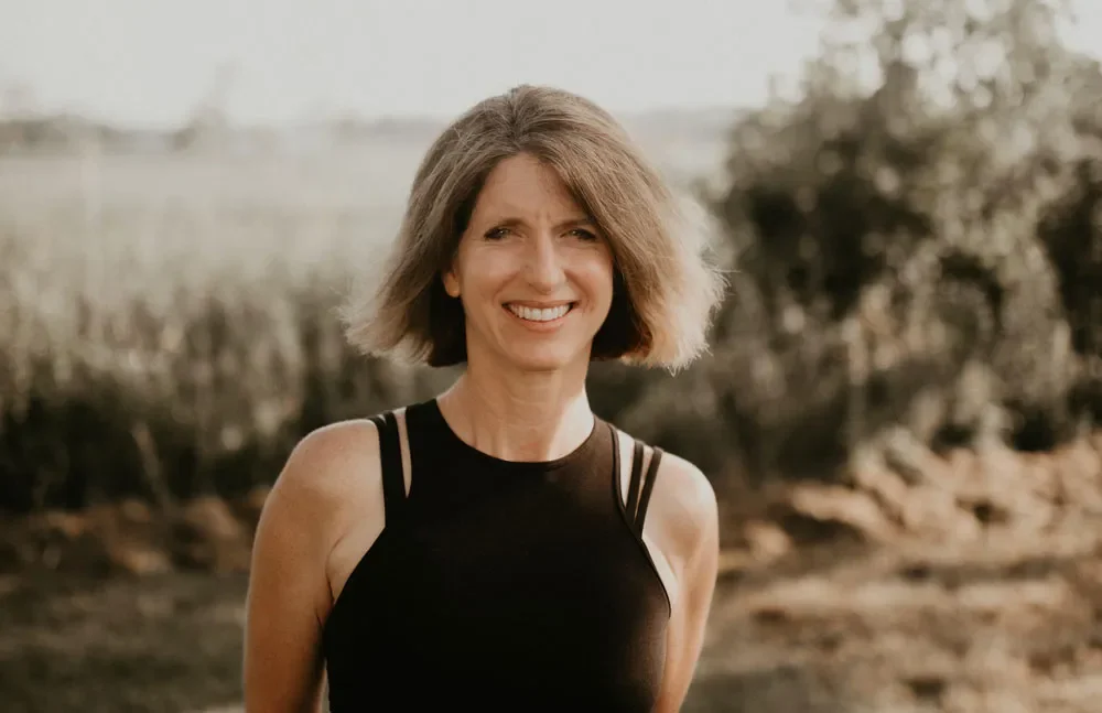 A smiling woman with shoulder-length light brown hair wearing a black tank top outdoors, with a blurry natural background.