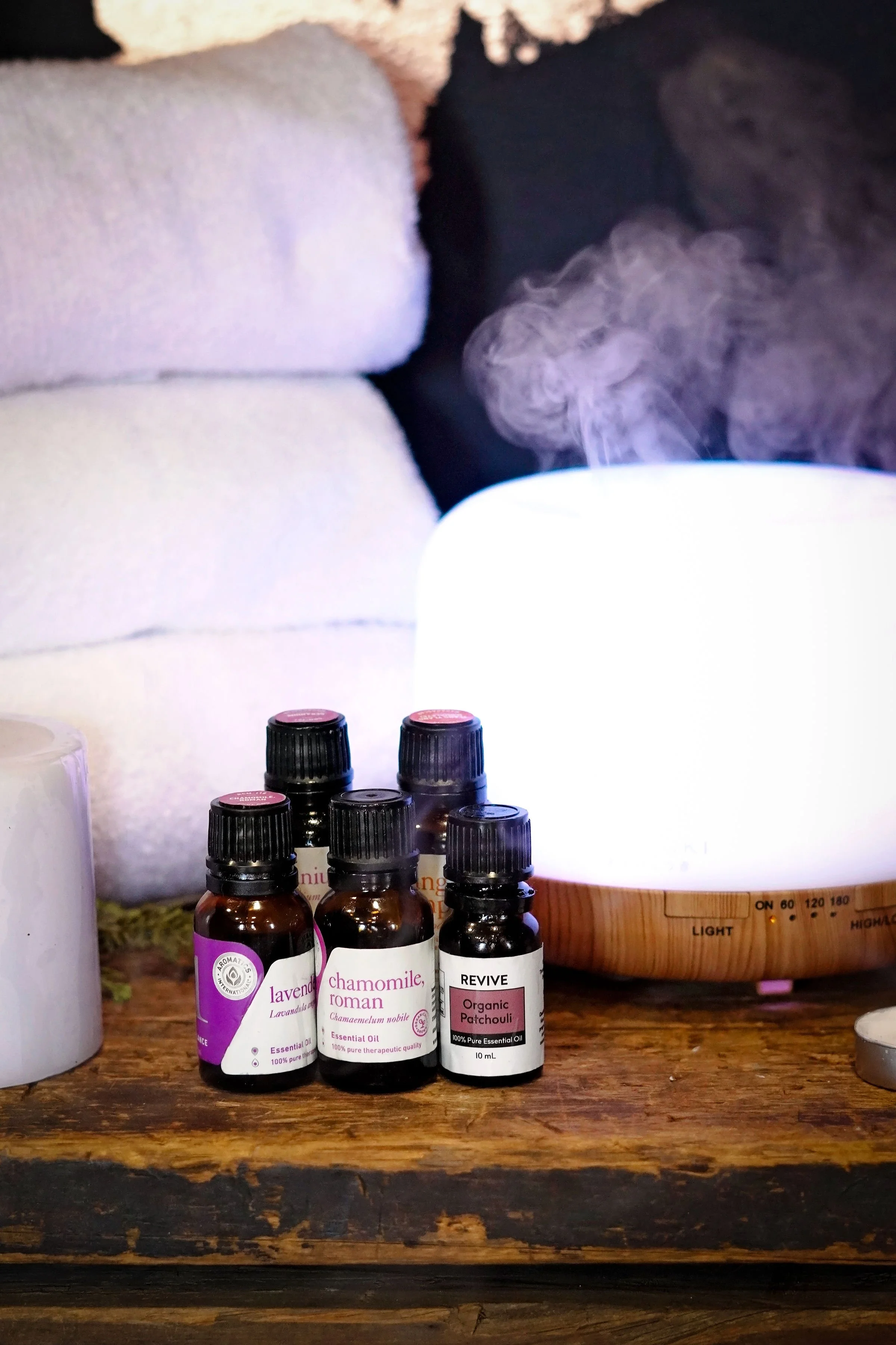 Several small bottles of essential oils with black caps and labels, placed on a rustic wood surface, in front of a white humidifier emitting vapor, with white towels in a cozy setting.