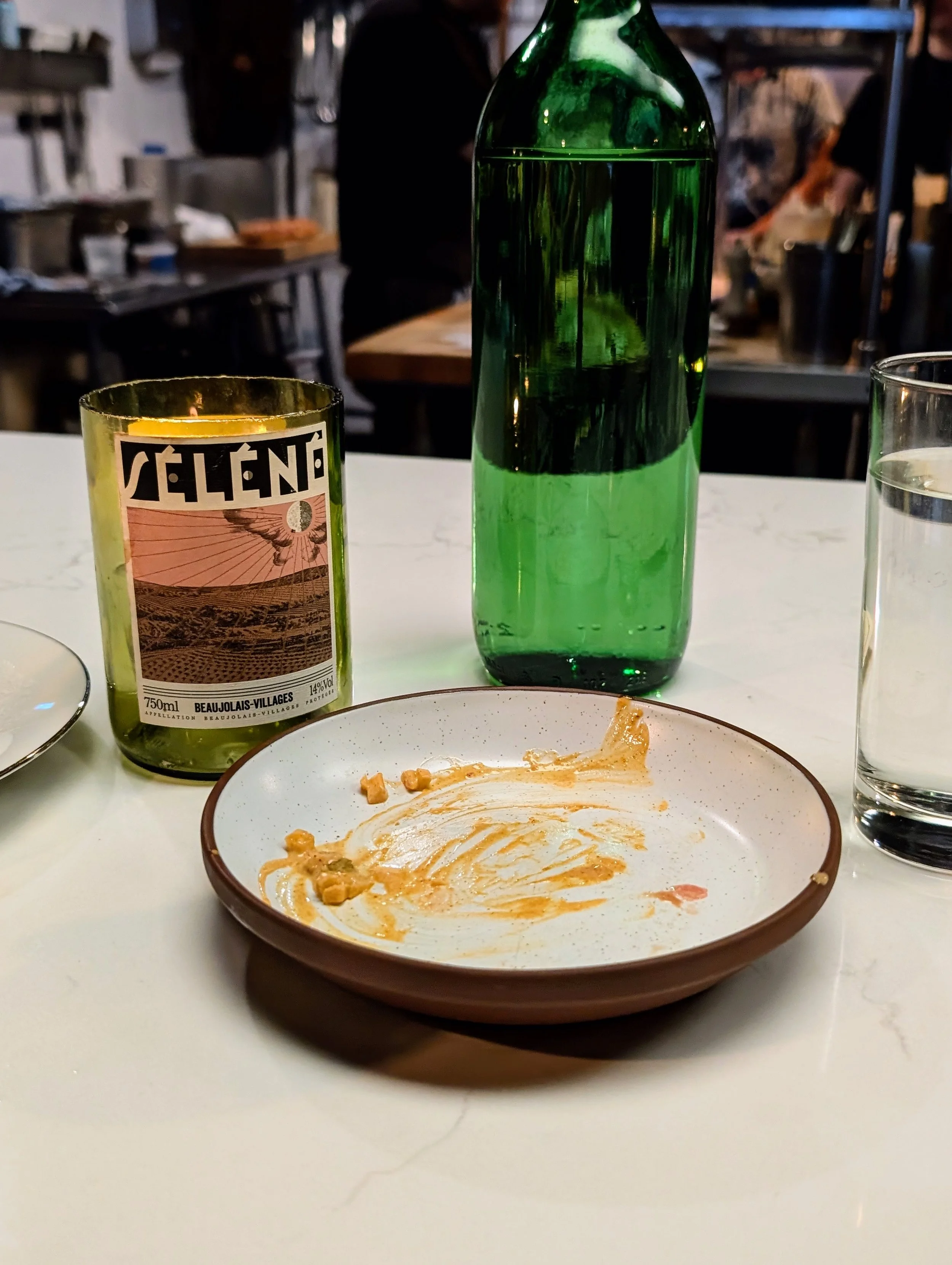 Empty plate with some remaining sauce and crumbs, a green bottle of water, a glass of water, and a candle on a white table at a restaurant.
