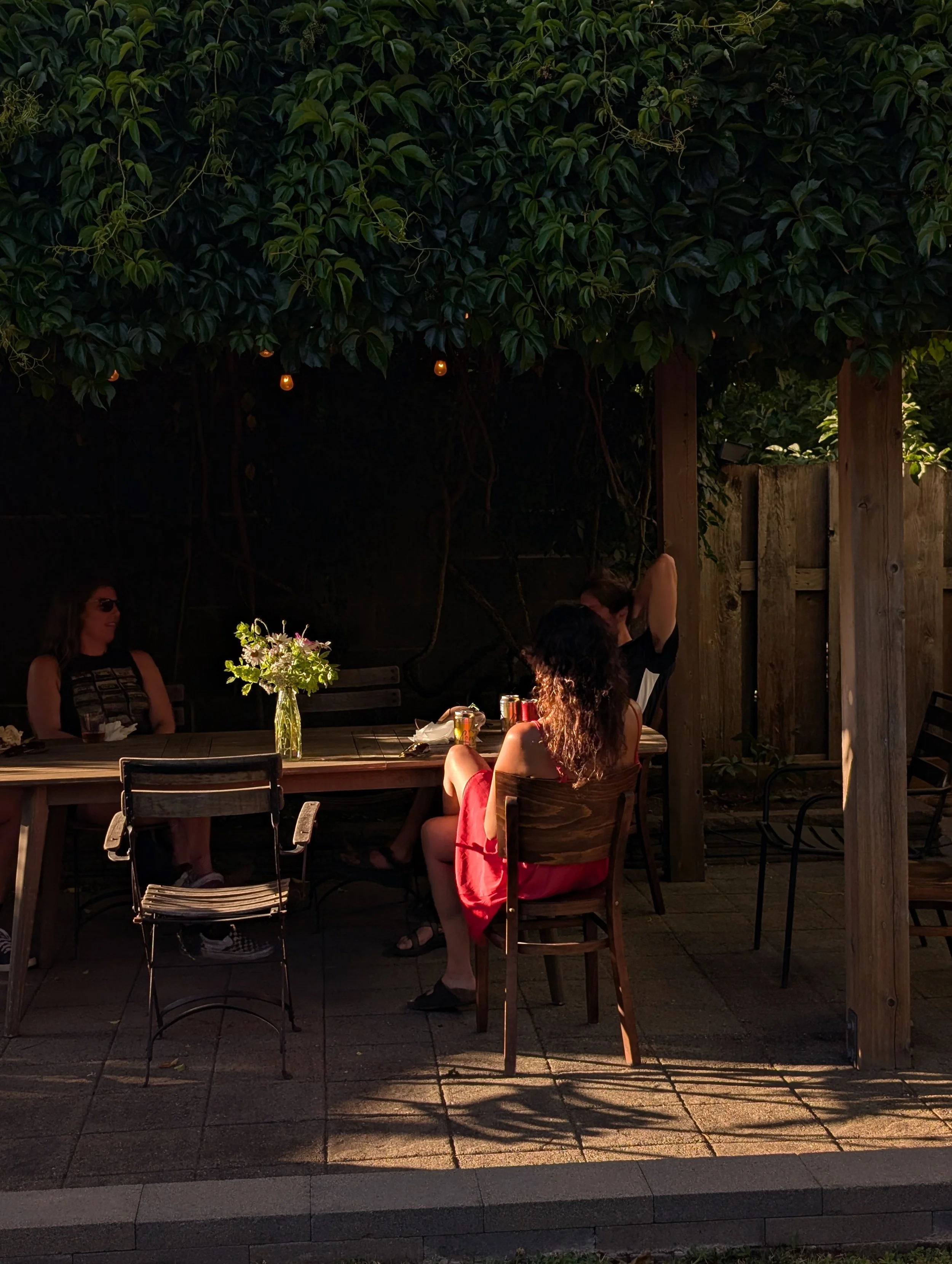 A group of four women sitting at an outdoor wooden table in a backyard patio, lit by sunlight, with a leafy hedge overhead and a flower vase in the center of the table.