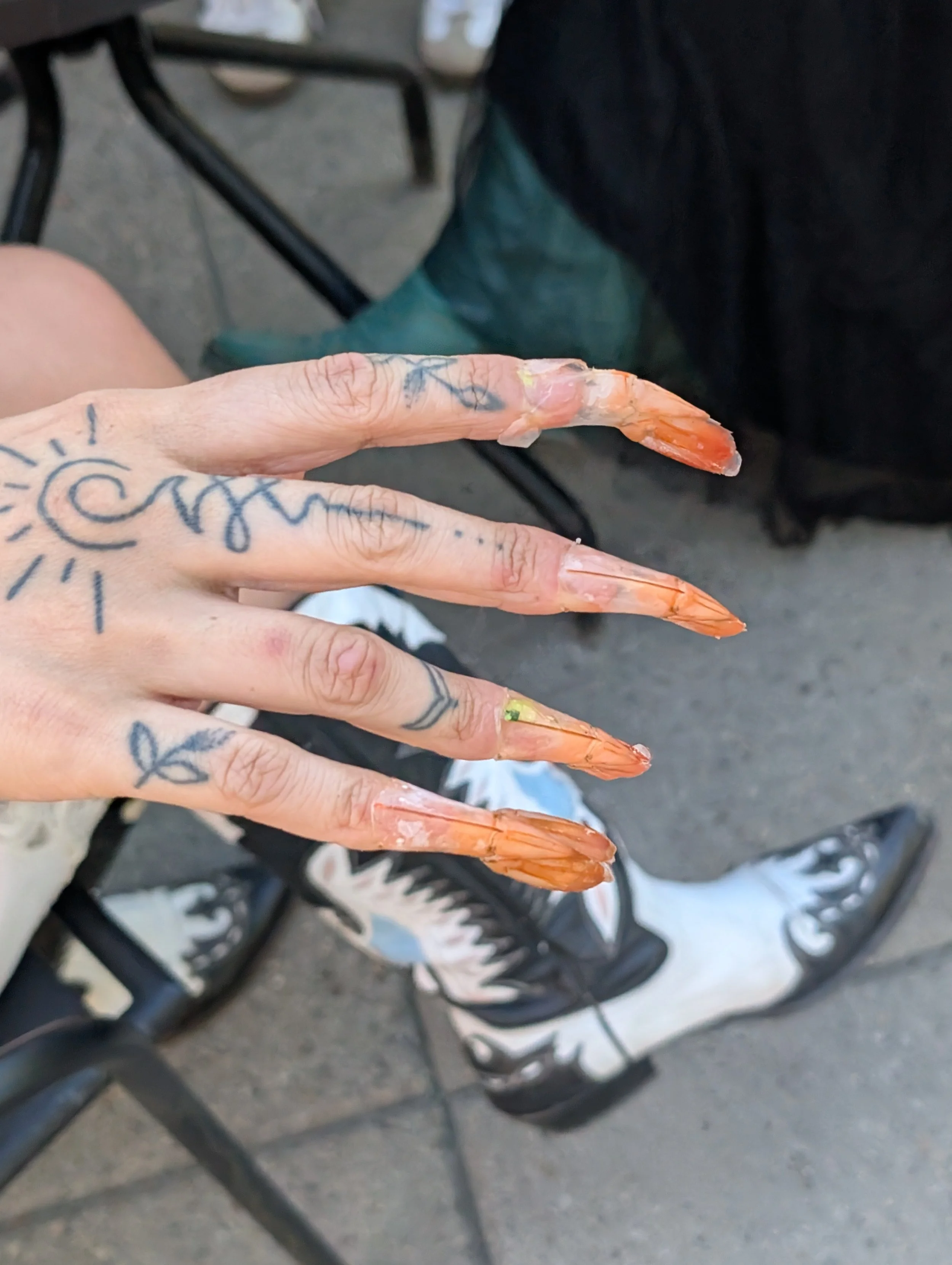Close-up of a person's hand with colorful tattoos, showing long, orange-tinted acrylic nails being painted or manipulated, with a blurred background of a chair, a person's leg in sneakers, and some outdoor flooring.