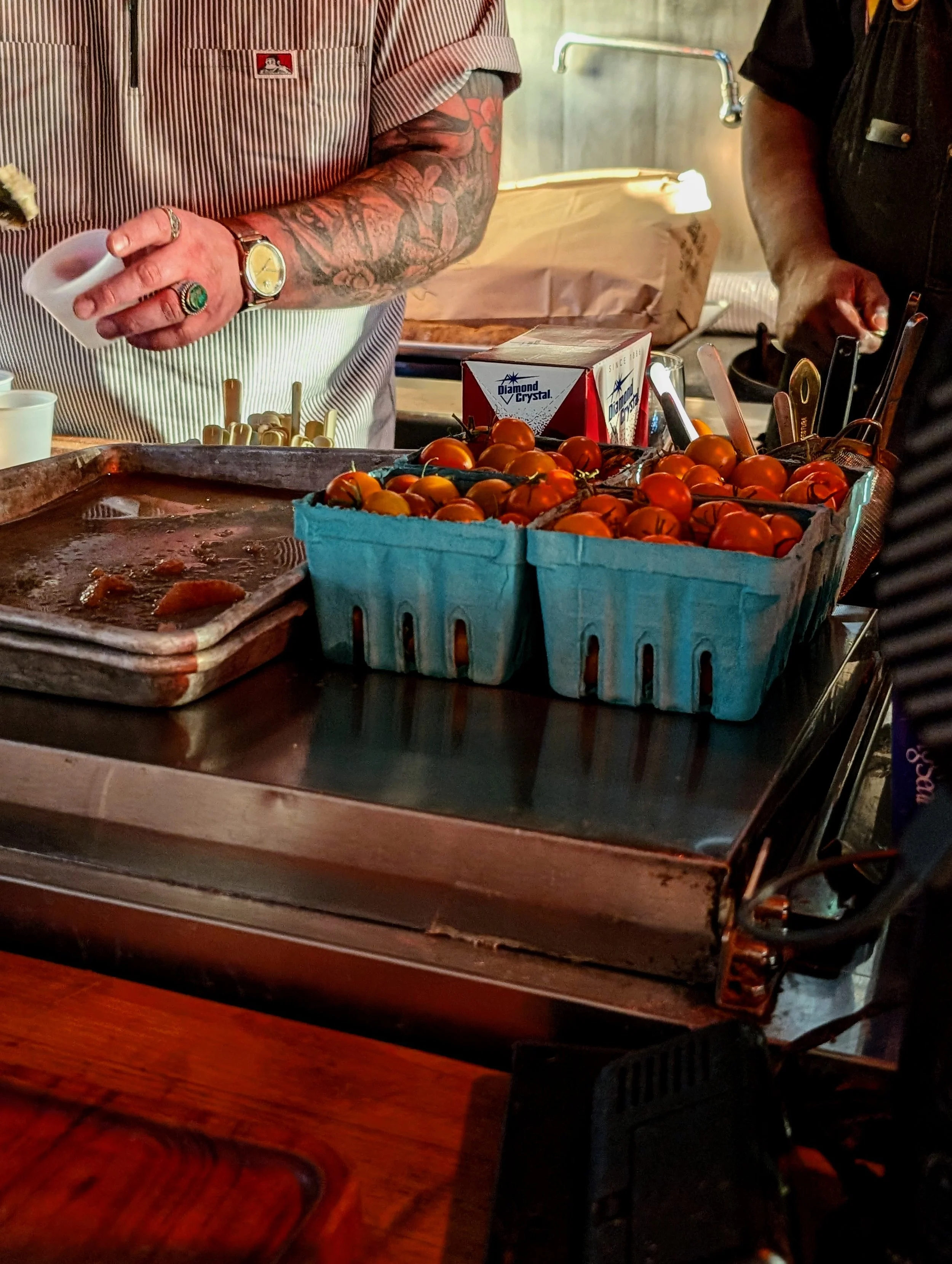 Cherry tomatoes in blue plastic baskets on a metal counter at a food stand, with two people preparing food behind it.
